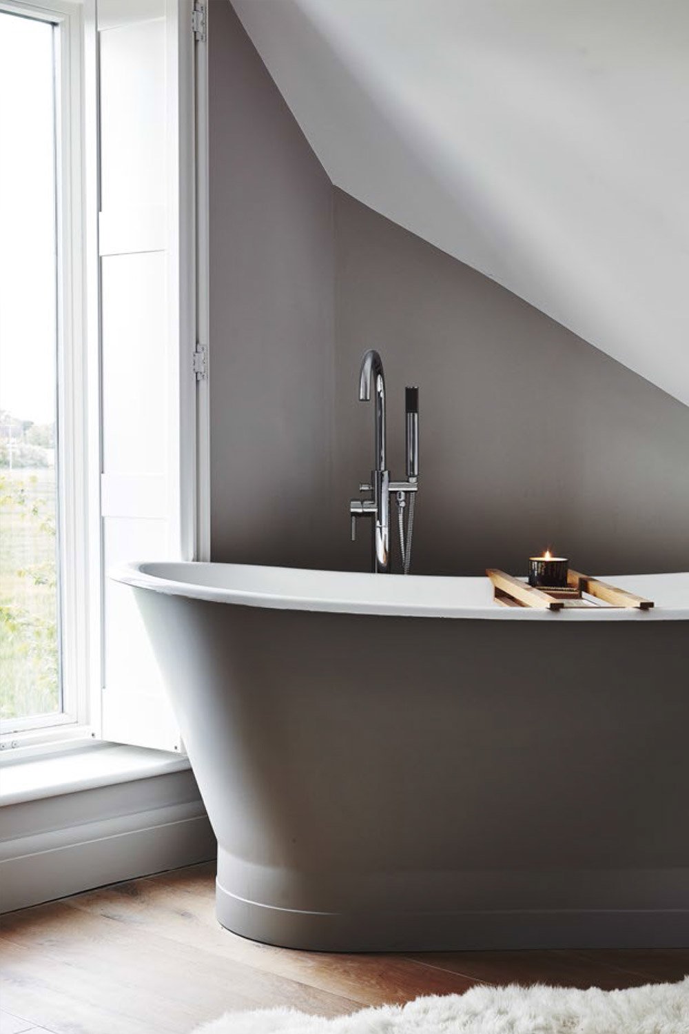 Modern freestanding bathtub with a gray exterior, situated next to a large window in a bathroom with gray and white walls, wood flooring, and a small black candle on a wooden tray.