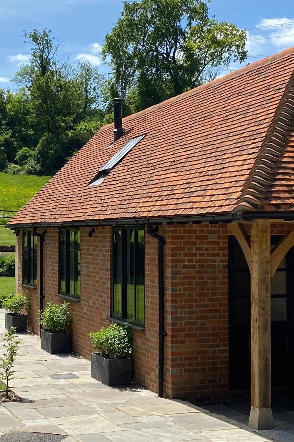 Side view of a brick house with black-framed windows, a tiled red roof, a chimney, and a skylight. The house has a wooden support beam at the entrance and landscaped potted plants along a stone walkway. In the background, there are green trees and a 