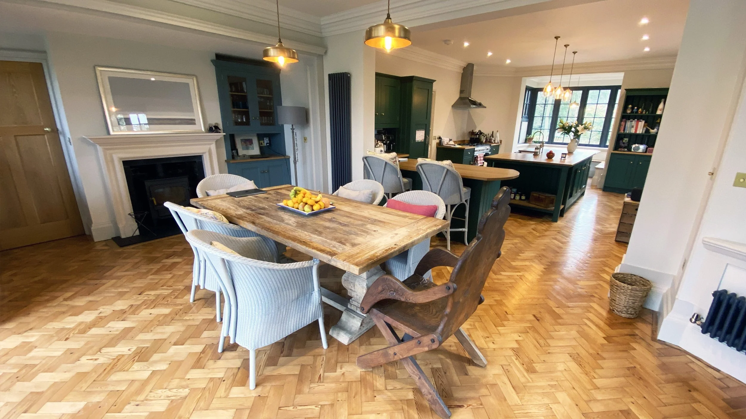 A cozy dining area in a kitchen with a rustic wooden table surrounded by various chairs. The room features hardwood flooring, a fireplace with a white mantel, dark green kitchen cabinets, and large windows letting in natural light. There are decorative lighting fixtures hanging from the ceiling, and a bowl of fruit on the table.