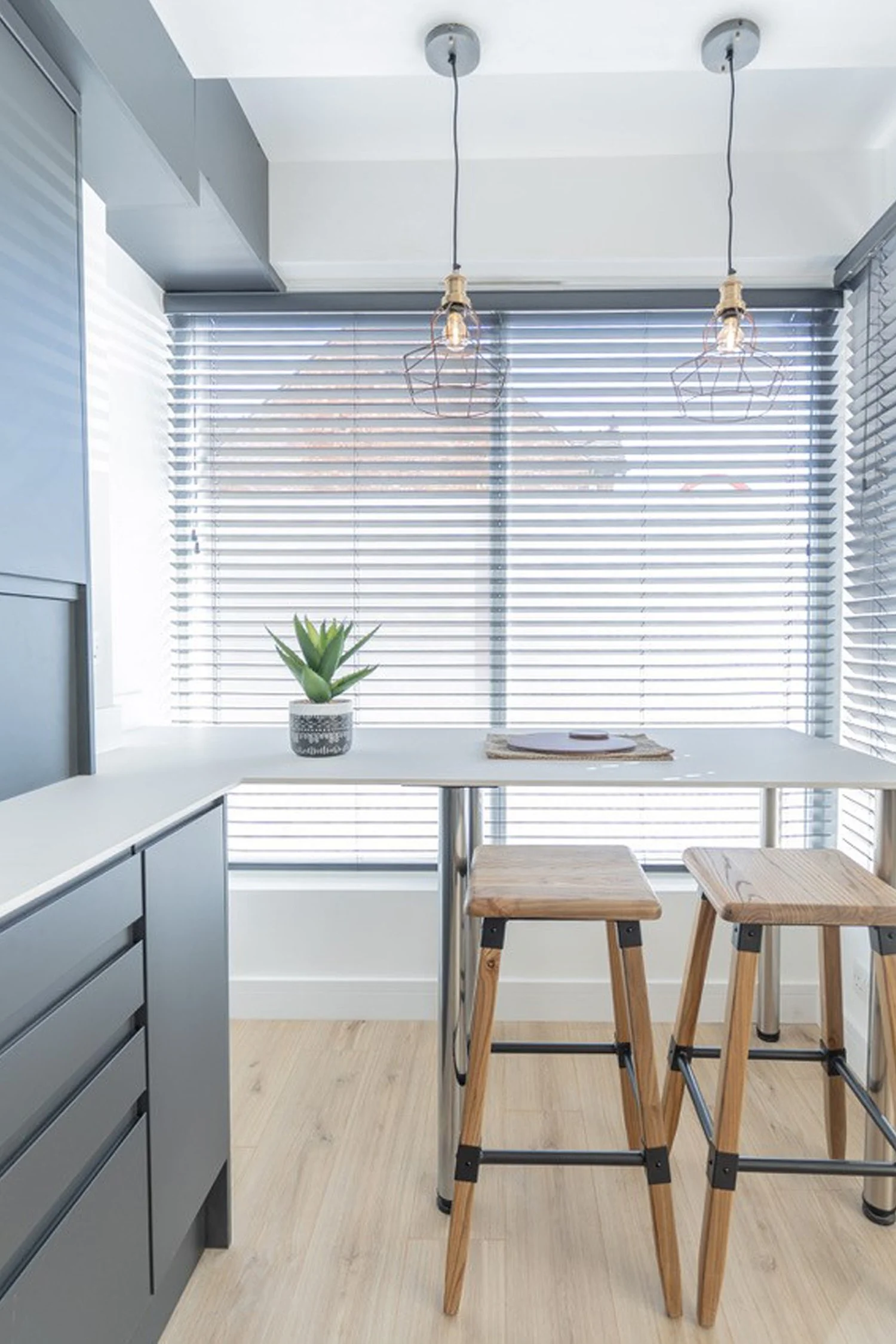 Modern kitchen with a white countertop, wooden stools, potted plant, and horizontal blinds on the window, hanging Edison-style light bulbs