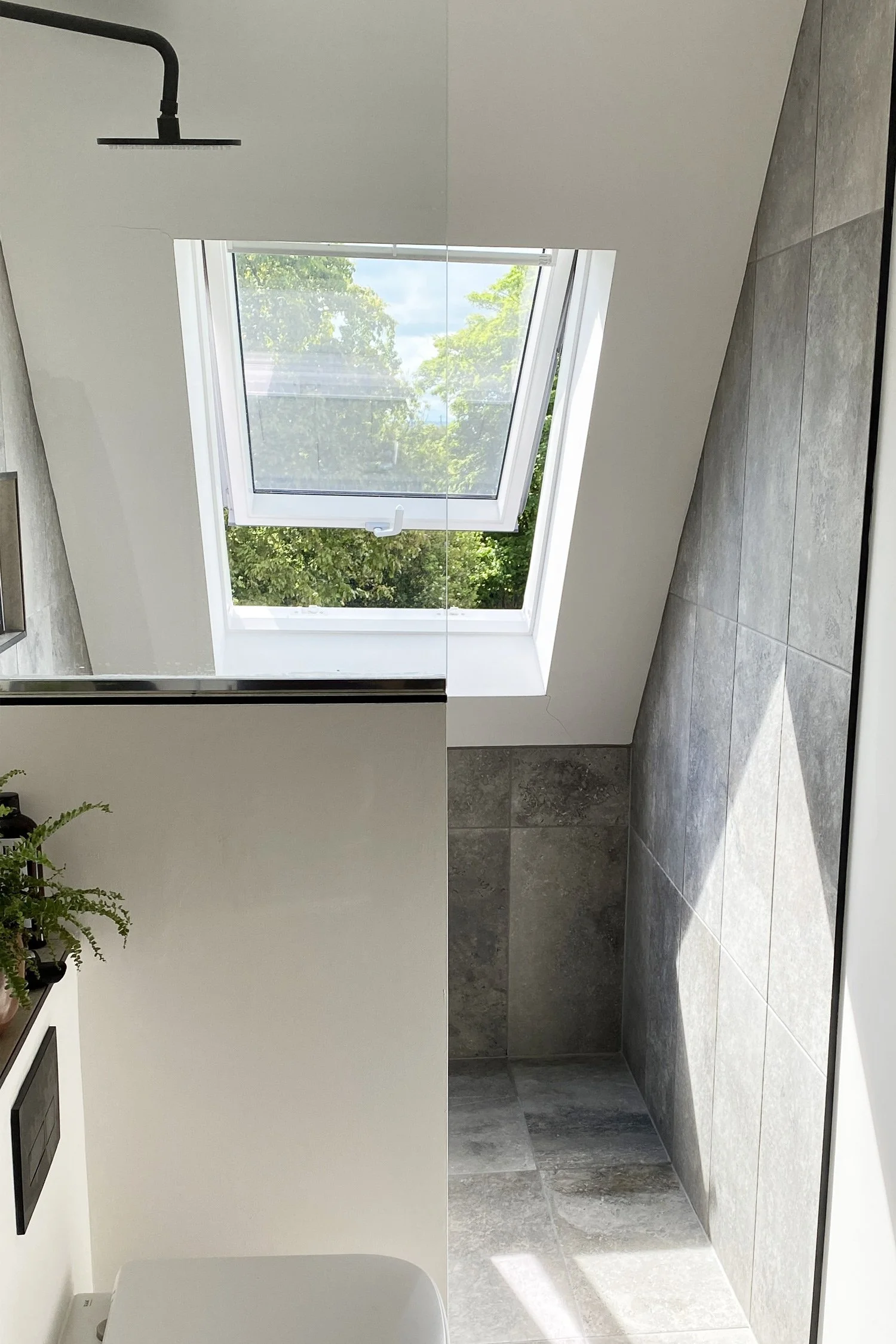A modern bathroom with a skylight window, gray tiled walls, and a white toilet. The ceiling is sloped, and there are green trees visible through the window.