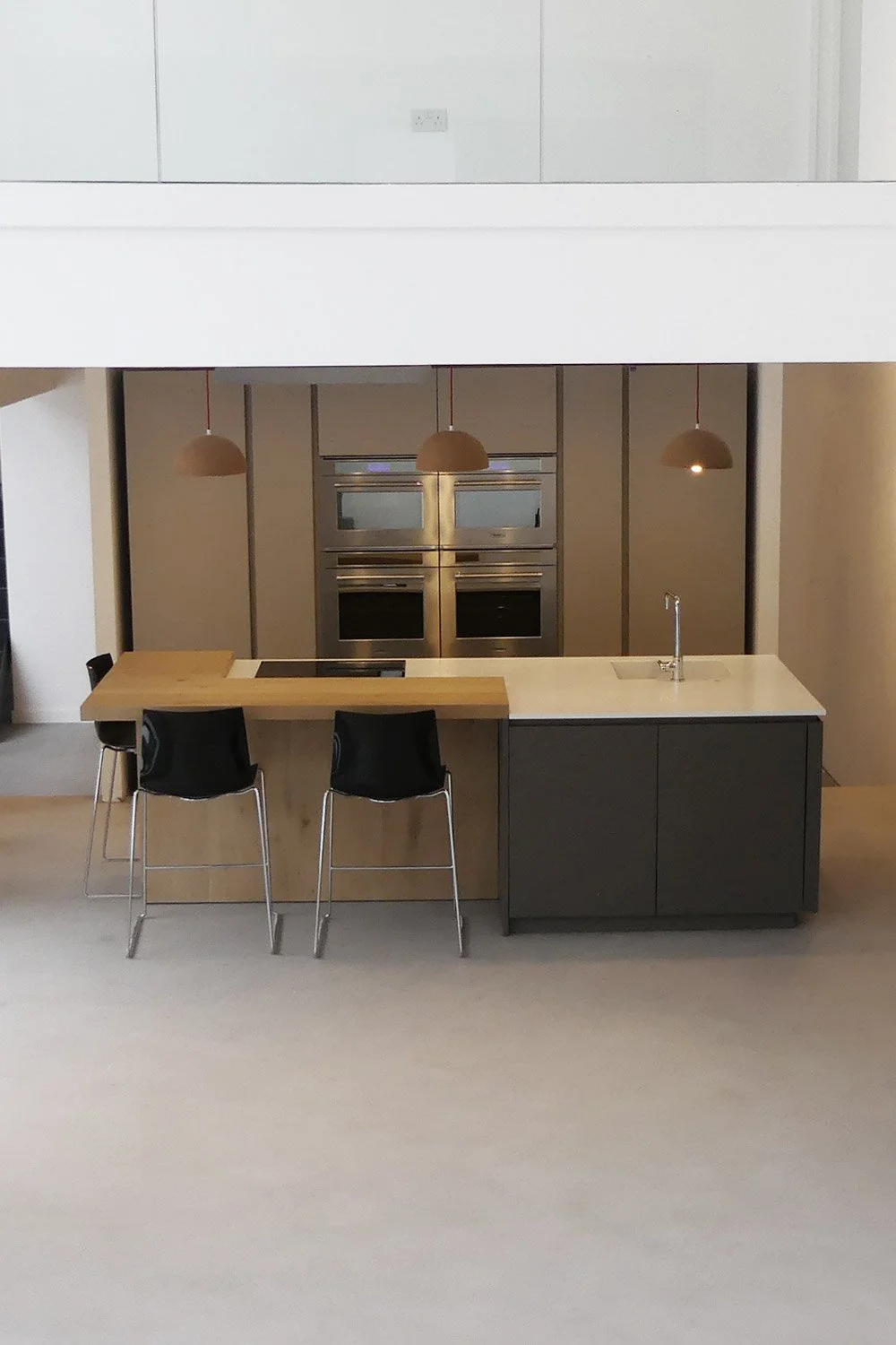 Modern kitchen with beige walls, black and wood cabinetry, three hanging pendant lights, black chairs at a two-tone kitchen island with sink, stainless steel ovens, and minimal decor.