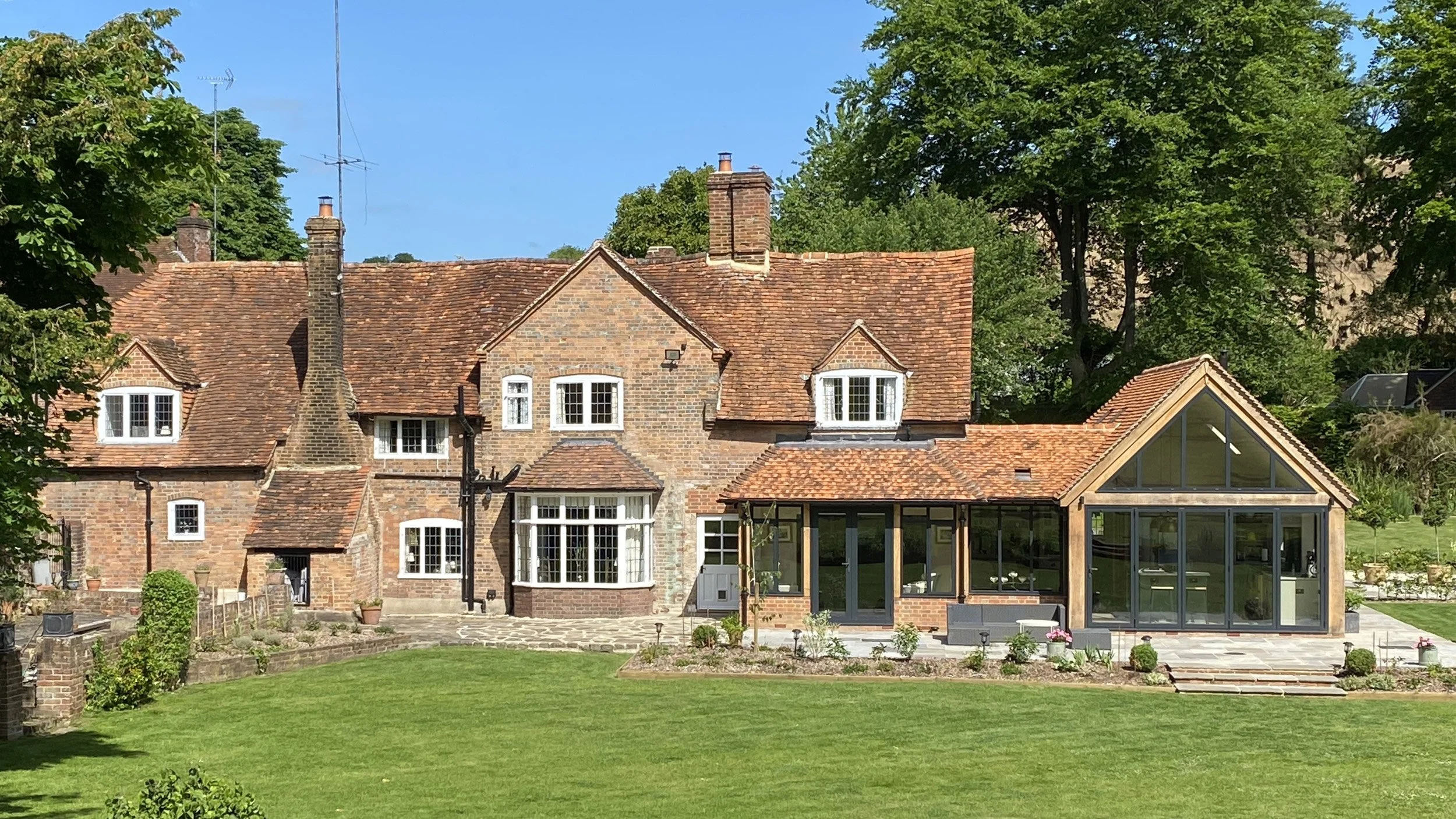 A large brick house with multiple gabled roofs, chimneys, and white-framed windows, with a modern glass extension and a patio in the backyard, surrounded by tall green trees and a well-maintained lawn.