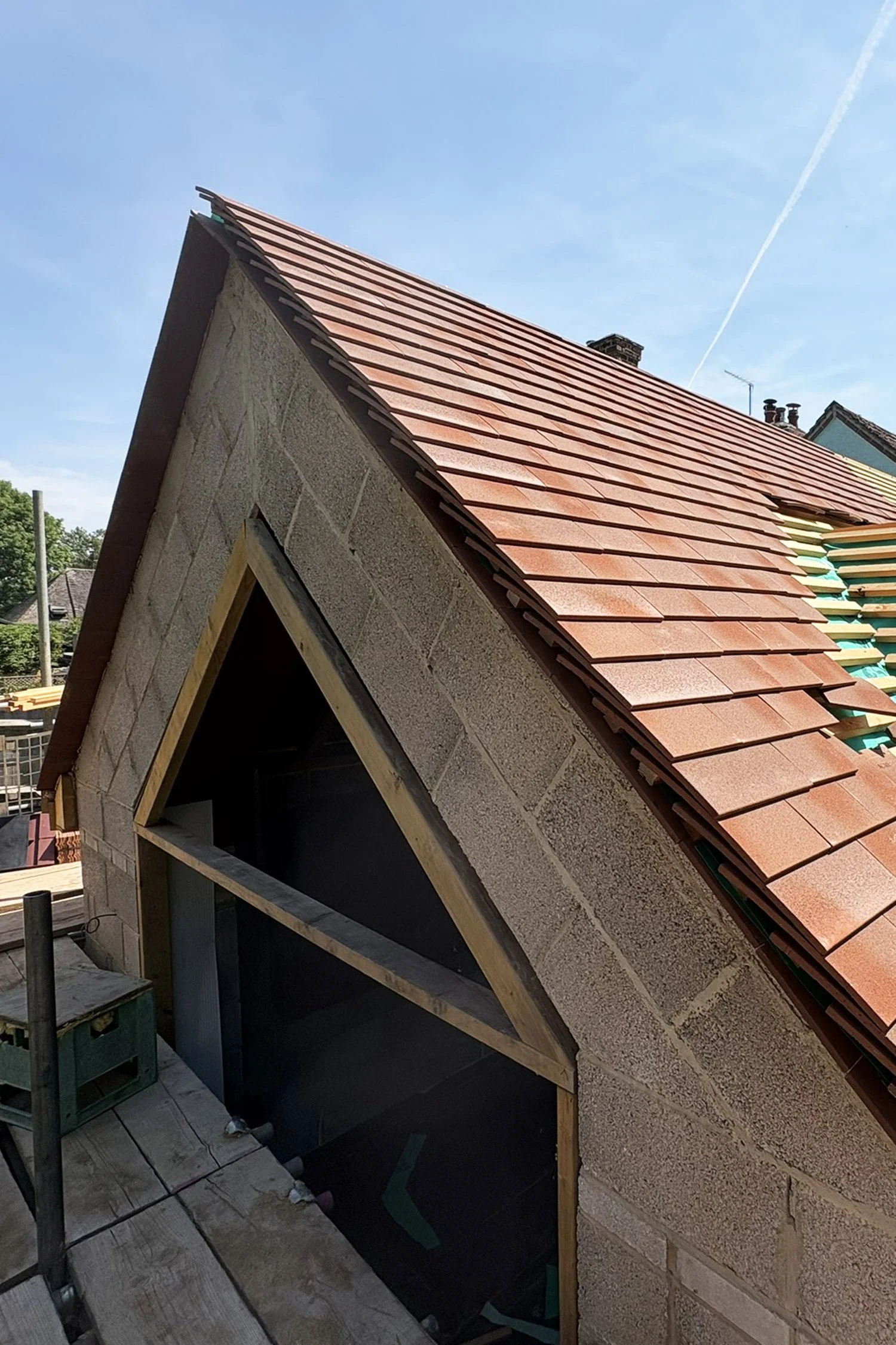 Roof under construction with red clay tiles being installed, showing a steep pitch and a chimney, part of a residential building.