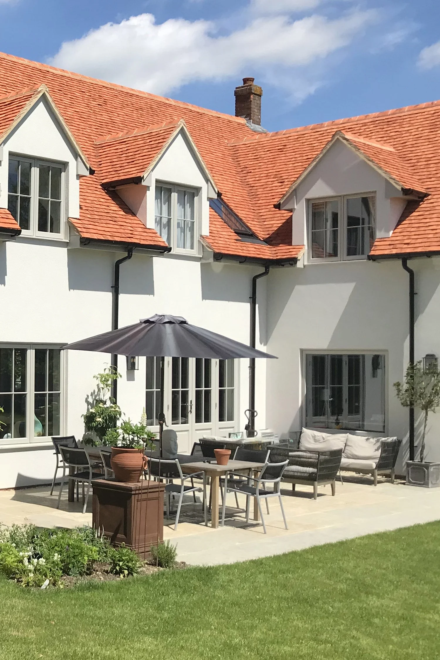 A backyard patio with outdoor furniture, a large patio umbrella, potted plants, and a well-maintained lawn, with a white house featuring a steep red-tiled roof and multiple dormer windows.