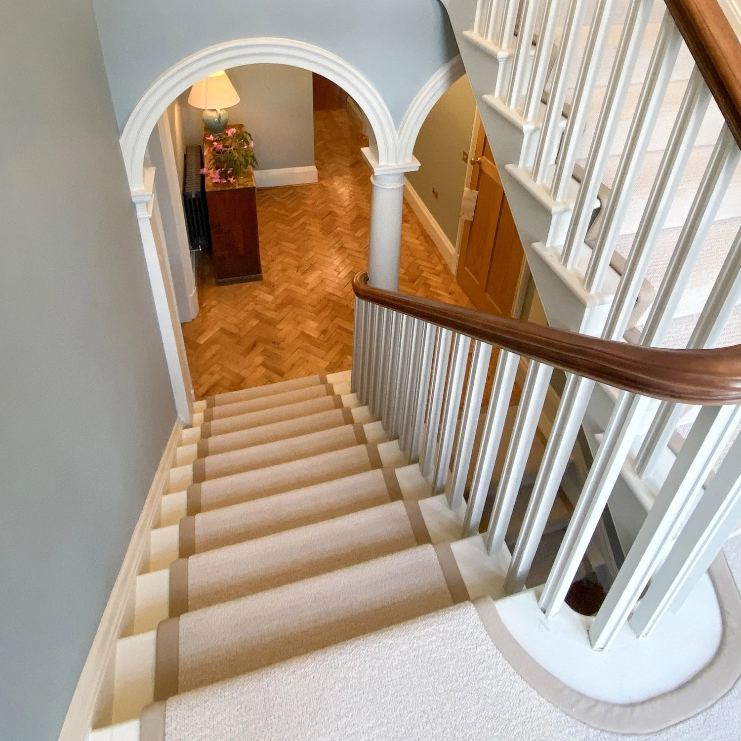 View of a staircase with beige carpet and a wooden handrail, looking down towards a hallway with wooden flooring, an arched doorway, and a table with a lamp and pink flowers.
