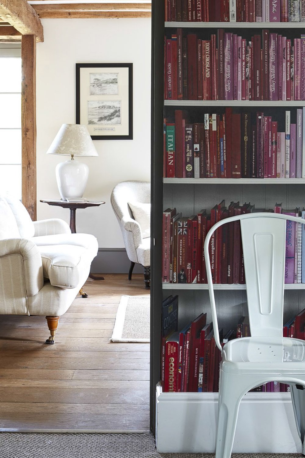 A cozy living room with white armchairs, a wooden side table with a white lamp, framed artwork on the wall, a wooden beam, and a bookshelf filled with red and purple books, a white chair, and a wooden floor.