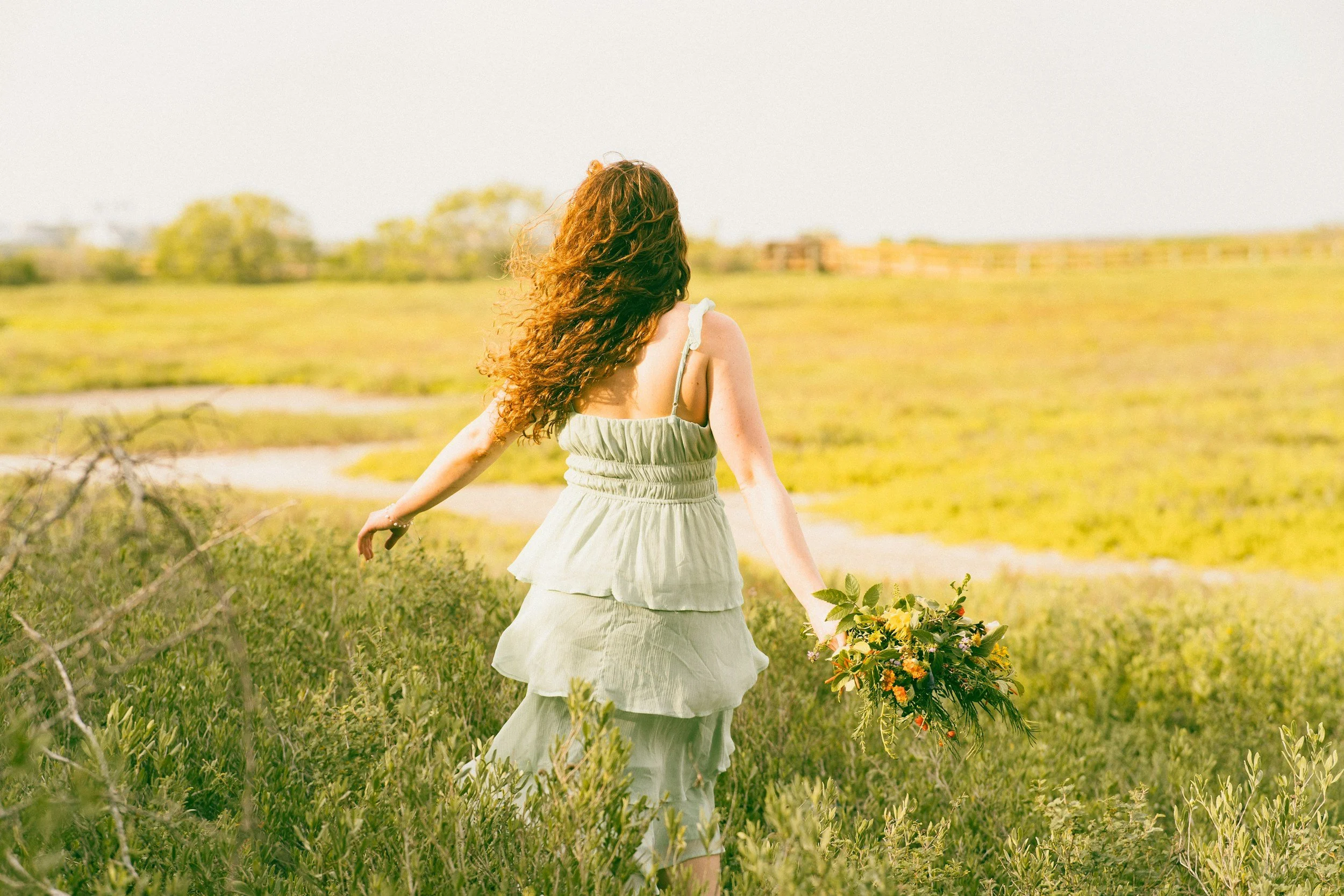A woman with long, curly red hair in a light green, tiered dress walking through a grassy field holding a bouquet of flowers.