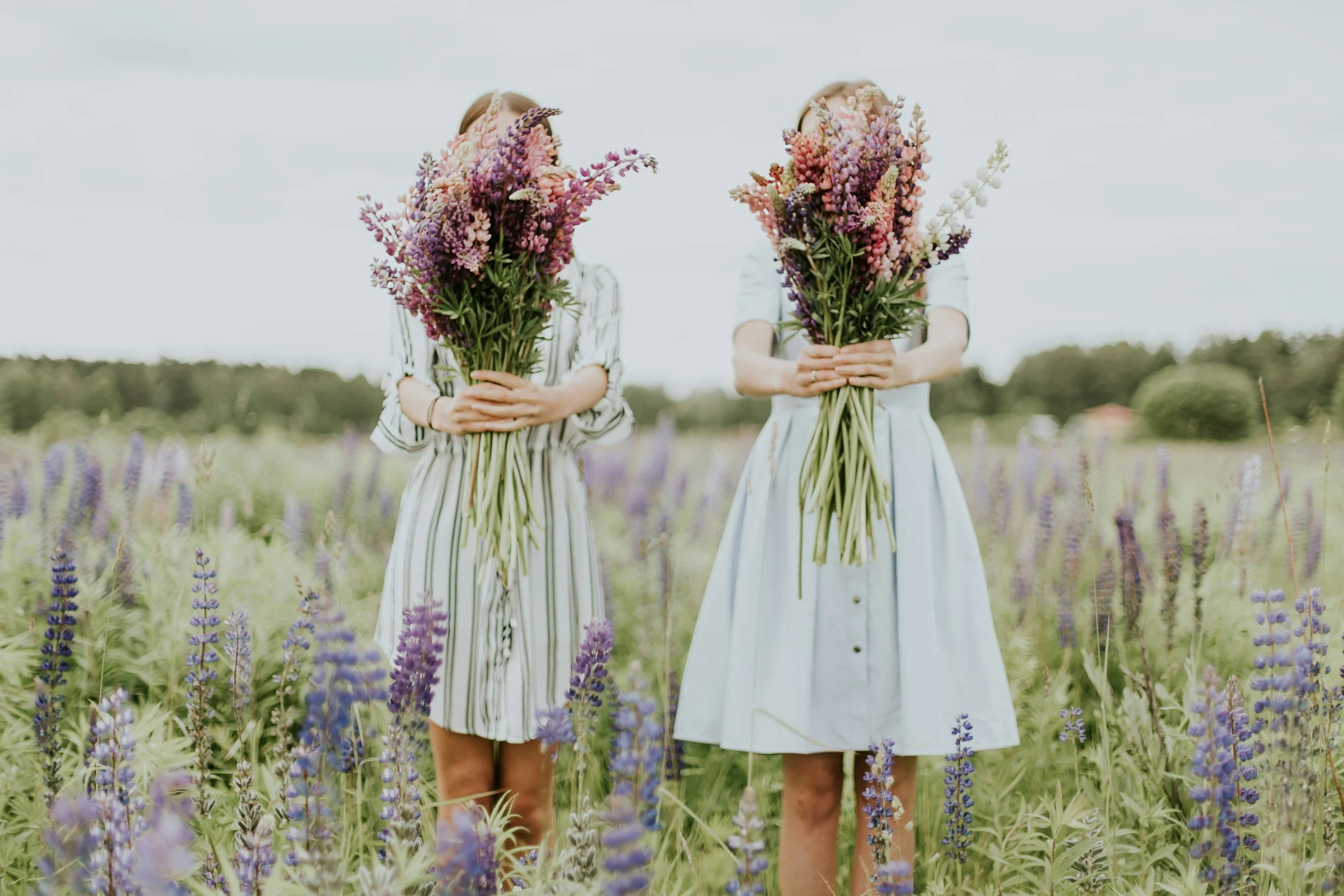 Two women standing in a field of purple and white flowers, each holding a large bouquet of pink, purple, and white flowers in front of their faces.