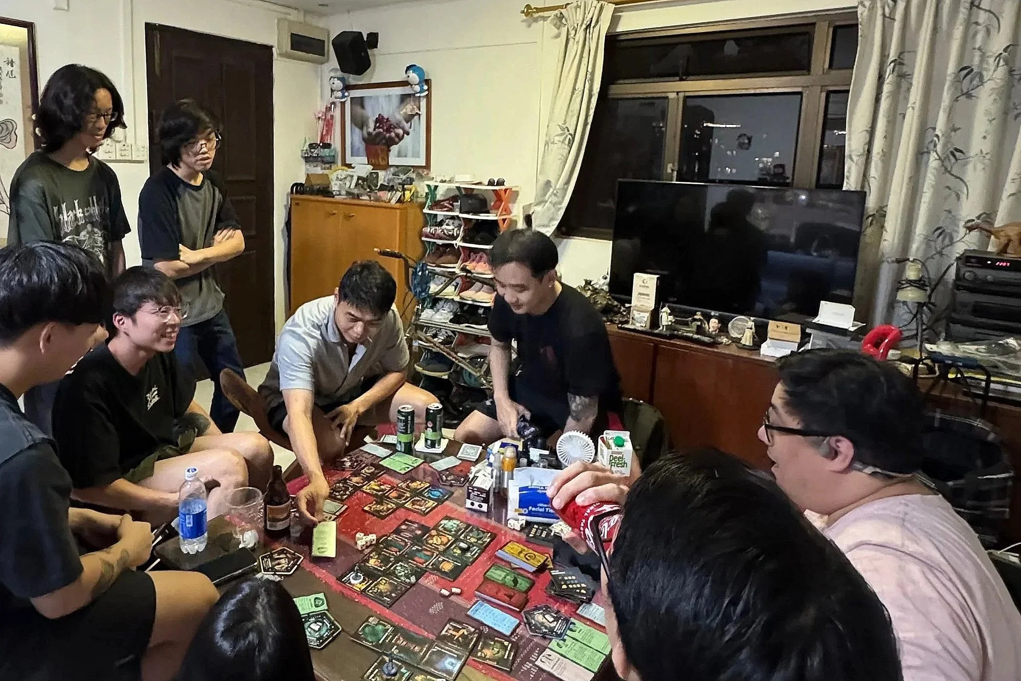 A group of people playing a board game (Betrayal at House on the Hill: 3rd Edition) around a table while the gamemasters watch.