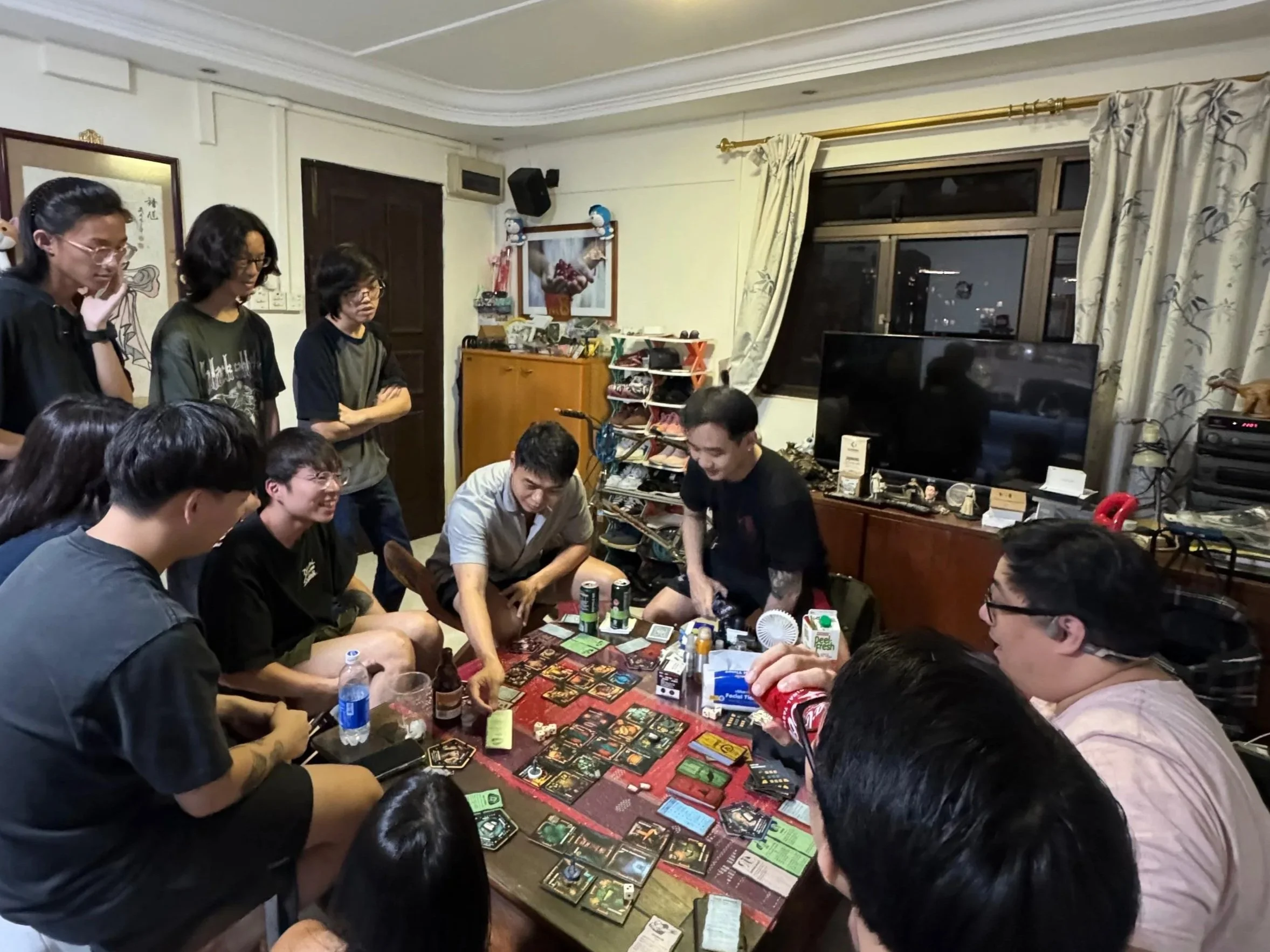 A group of people playing a board game (Betrayal at House on the Hill: 3rd Edition) around a table while the gamemasters watch.