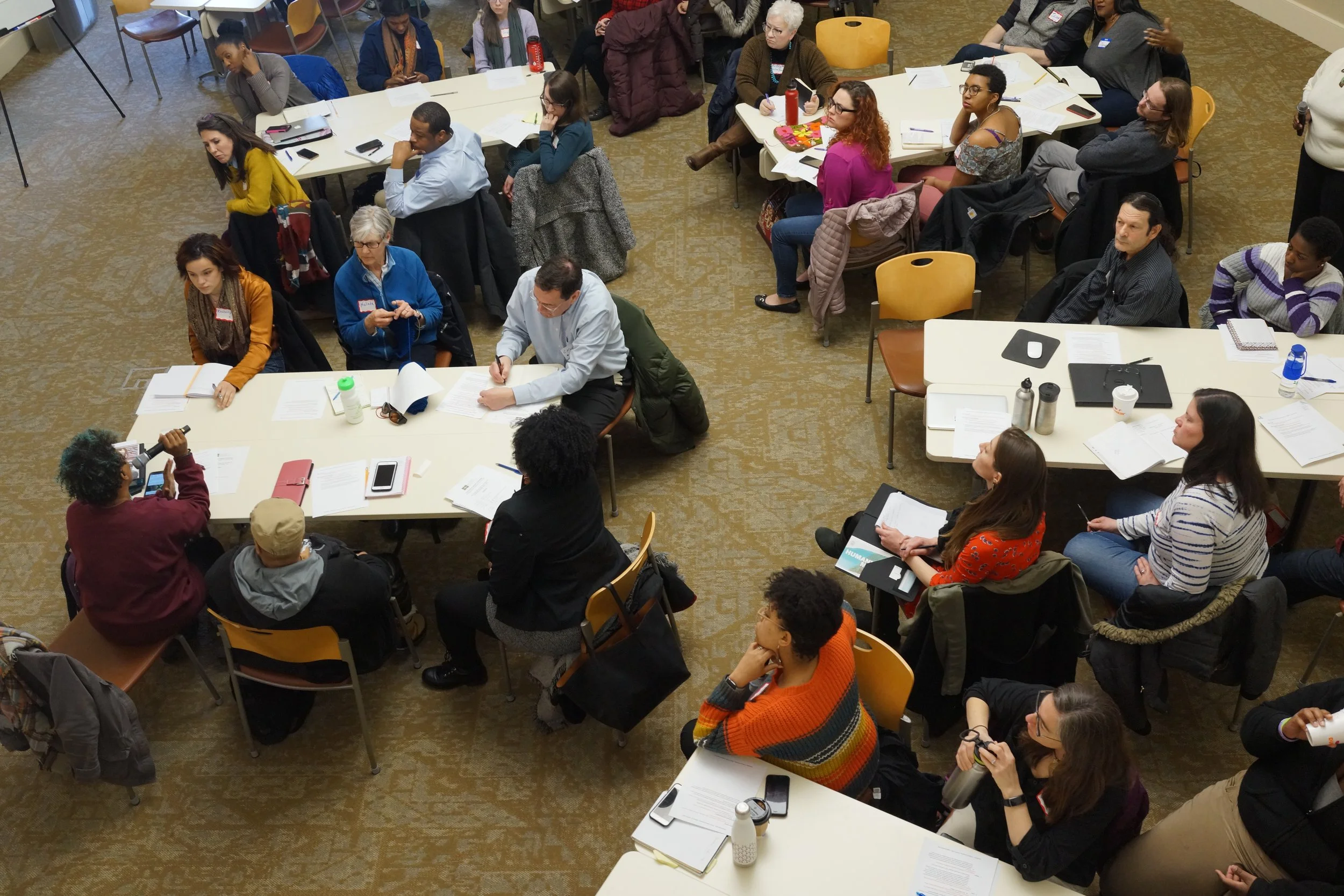 An overhead view of a diverse group of people seated around several large tables in a conference or workshop setting. Some individuals are taking notes, listening attentively, or engaged in discussions, with a few standing or speaking. The room has a