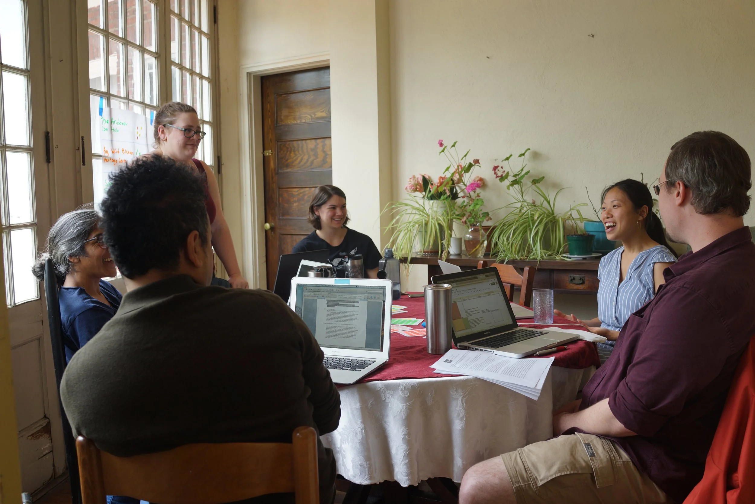 A group of people sitting around a table during a meeting or discussion, with laptops, papers, and drinks, in a room with large windows and potted plants.