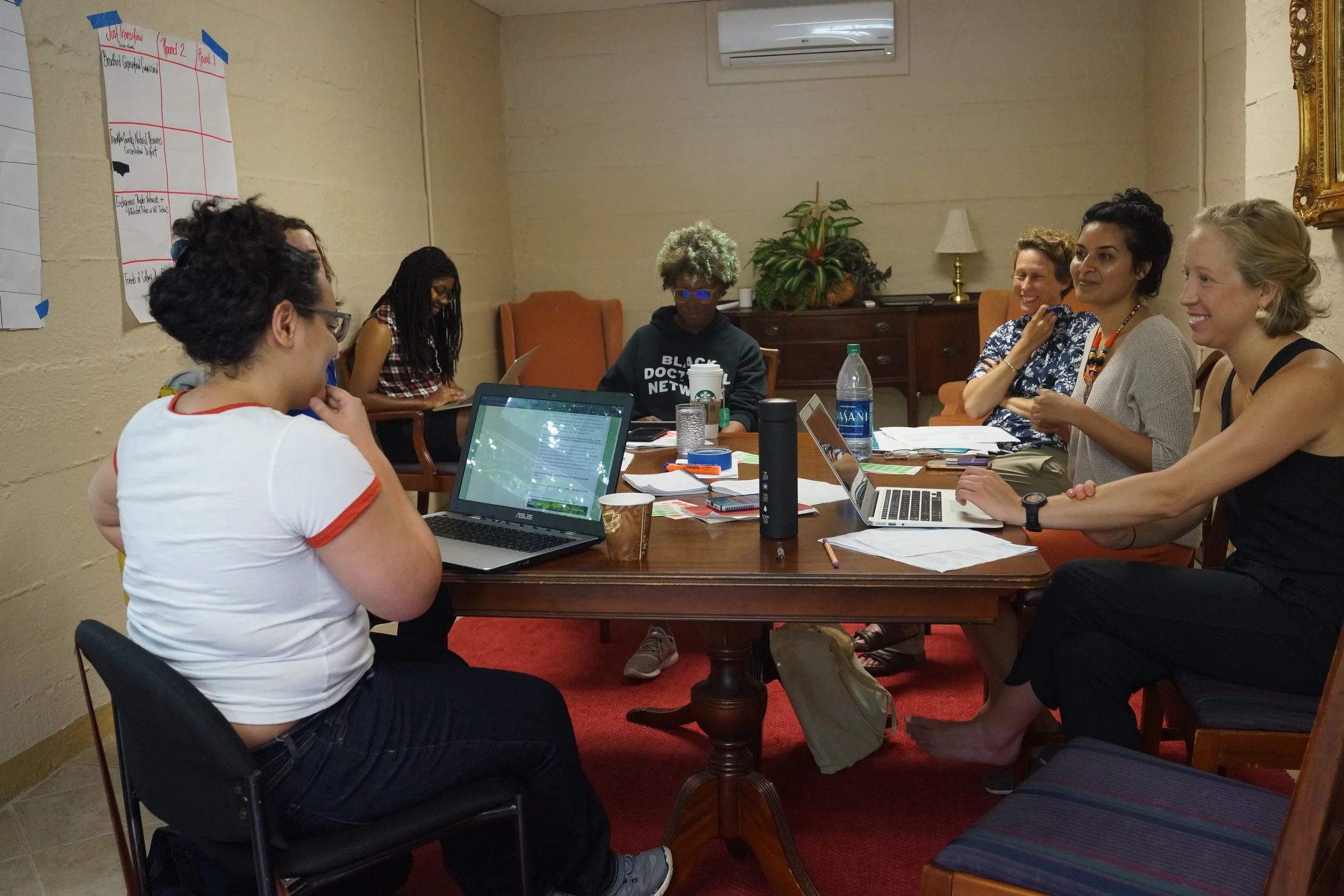 A diverse group of women sitting around a wooden table in a meeting room, smiling and engaging in discussion. There are laptops, notebooks, and water bottles on the table, with indoor plants and a framed mirror on the wall in the background.