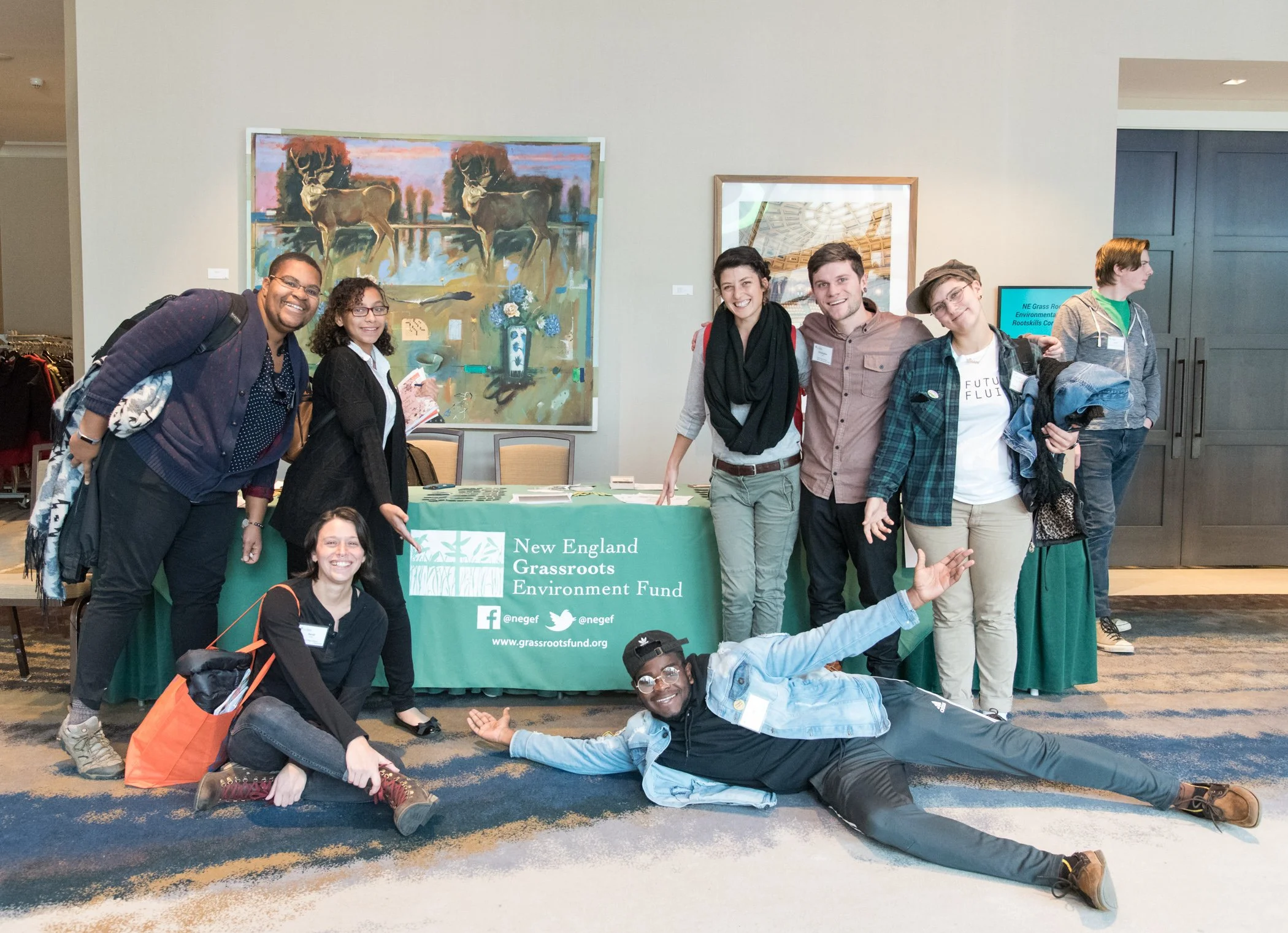 A group of people at an event, standing behind a table with a green tablecloth that reads 'New England Grassroots Environment Fund,' with some people sitting or lying in front. The background has large artwork and a television screen.