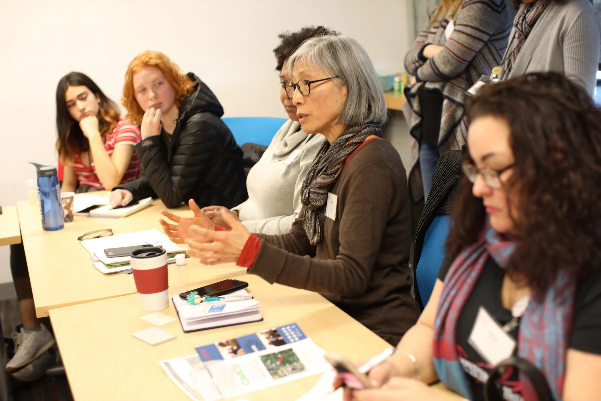 A group of women sitting at a table in a meeting or workshop, listening and engaging in discussion. The table has notebooks, magazines, and a drink cup.