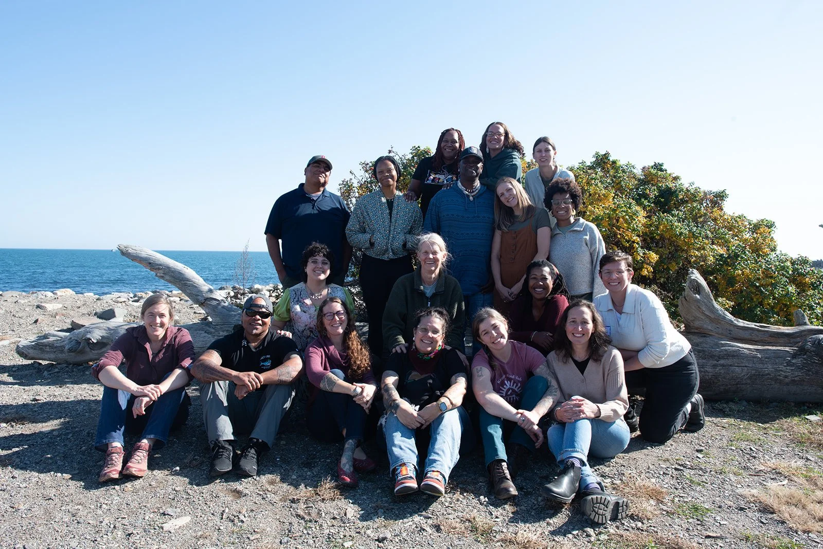 Group of diverse people smiling and posing together on a rocky beach with driftwood, trees, and ocean in the background.
