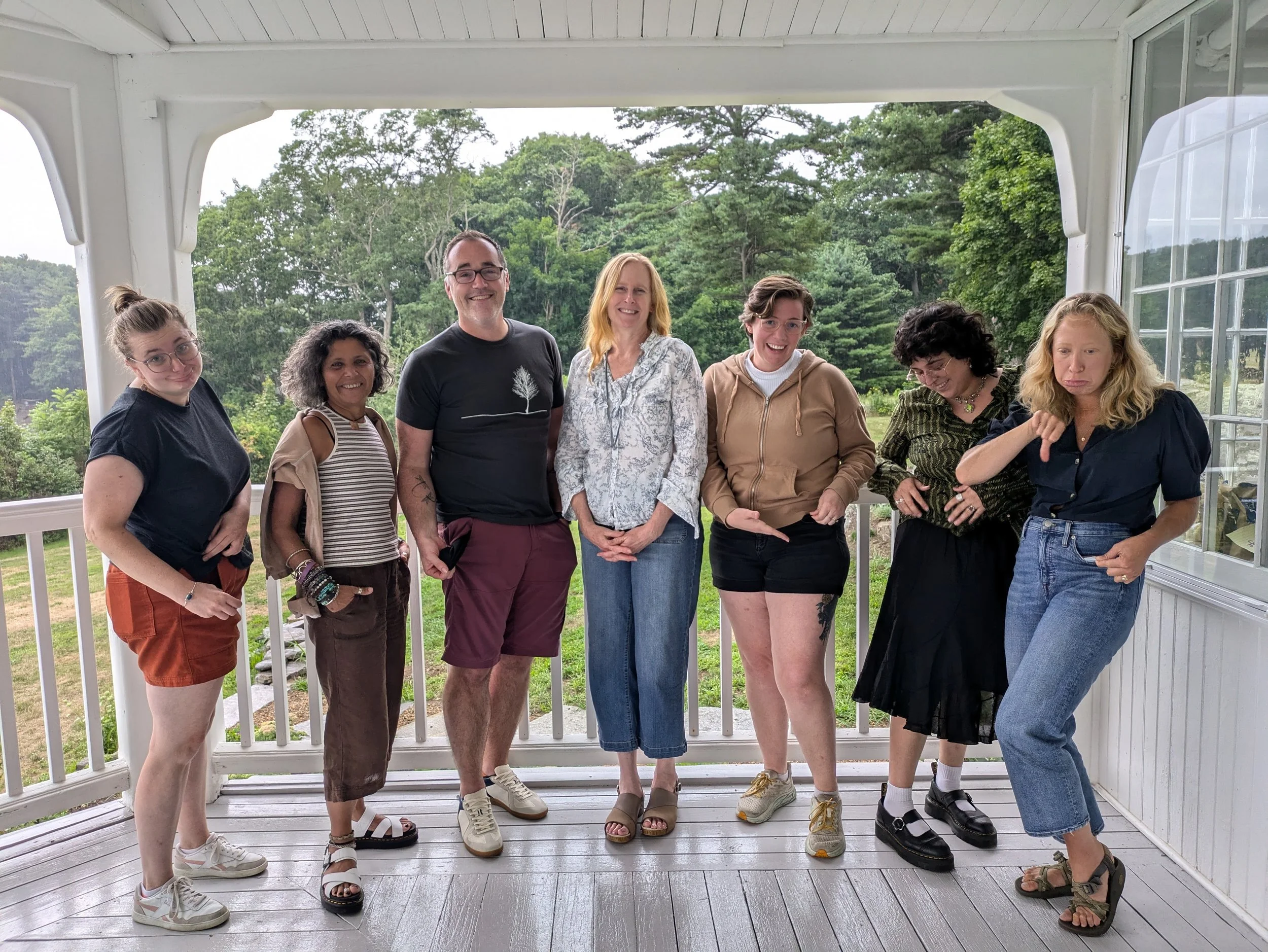 A group of seven people standing on a porch with a railing, with a green, wooded area in the background. They are smiling and wearing casual summer clothing.