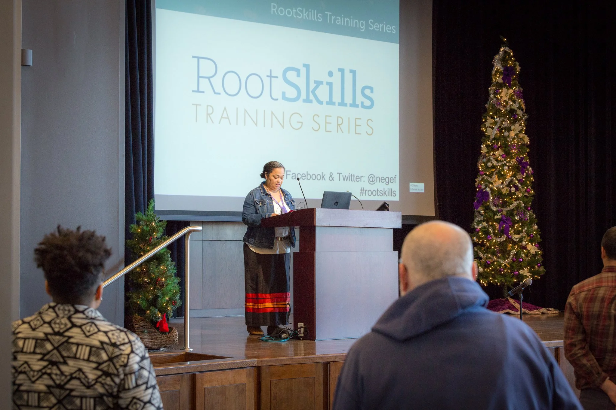 A woman standing at a podium giving a presentation during a RootsSkills training series, with a large projected slide behind her and a decorated Christmas tree to the right.
