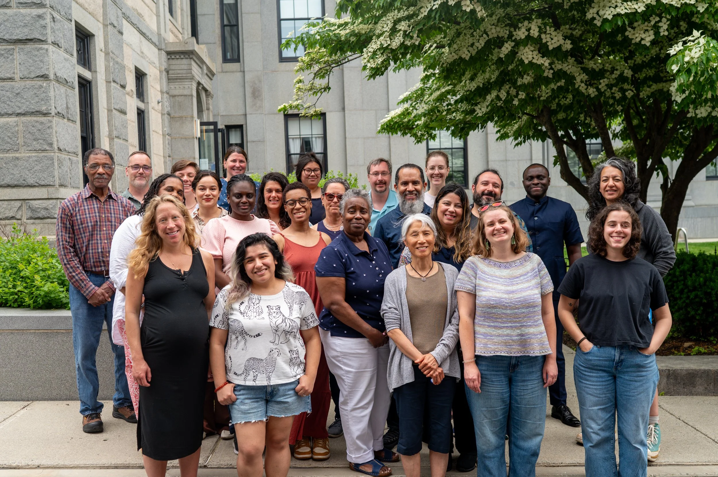A diverse group of people standing together outdoors on steps, in front of a building with large windows and trees, smiling at the camera.