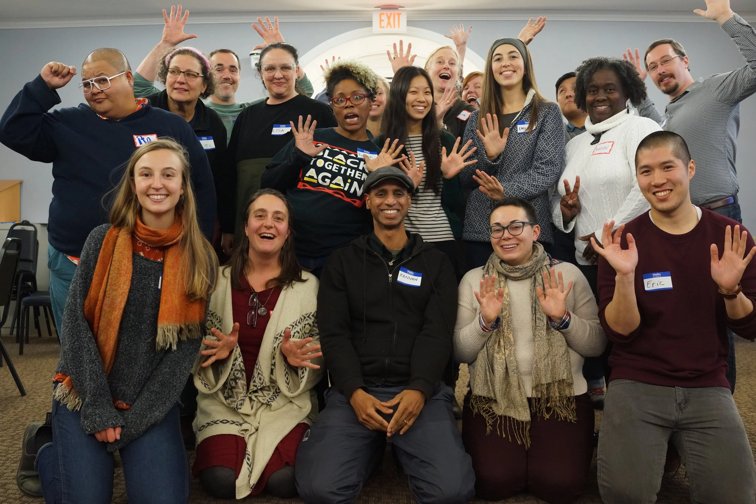 A diverse group of people gathered indoors, smiling and waving at the camera, with some wearing name tags, in a room with an exit sign overhead.