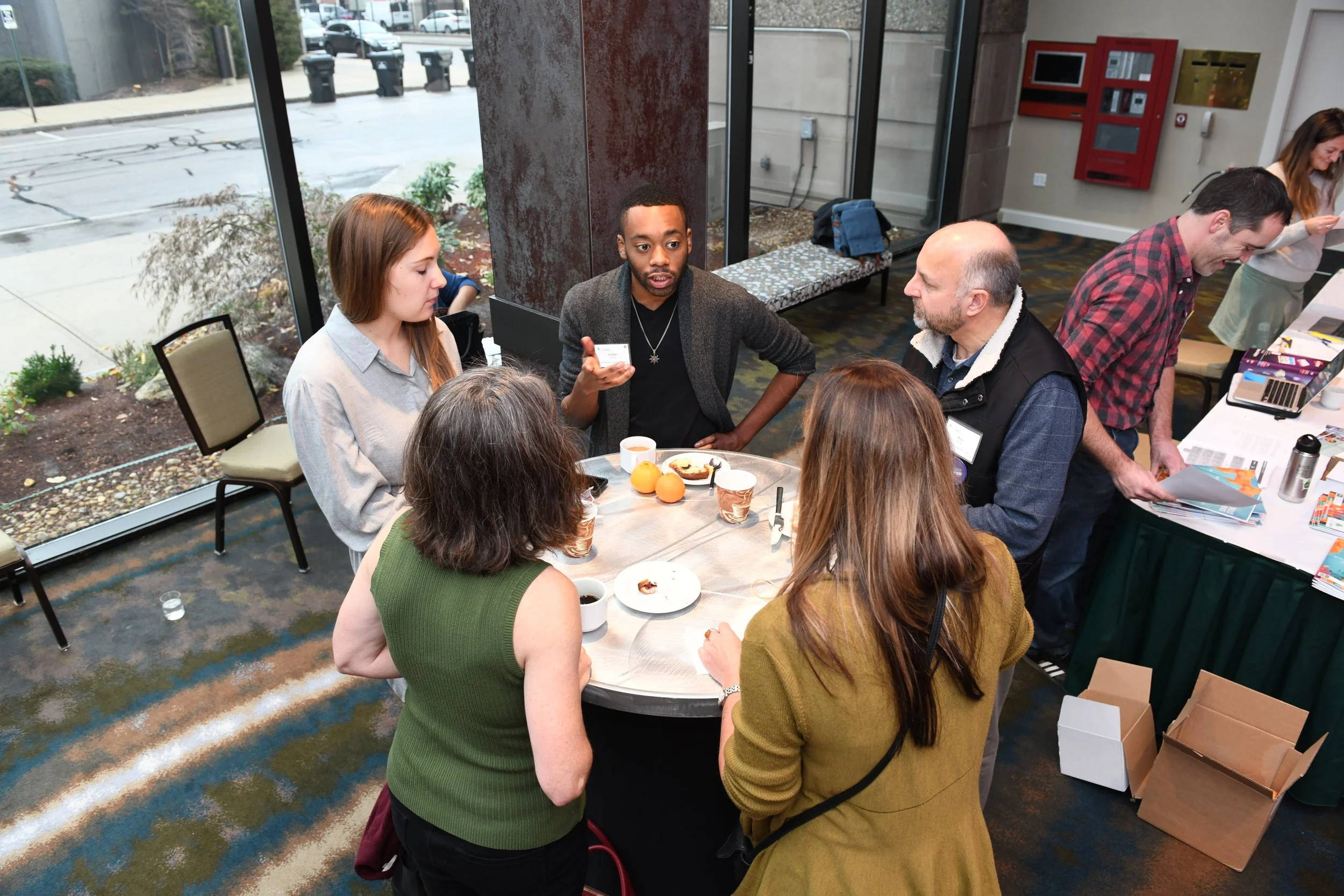 A group of diverse people gathered around a circular table having a discussion in a conference room with large glass windows showing an outdoor parking lot. There is food, coffee cups, and papers on the table, with some individuals explaining or list