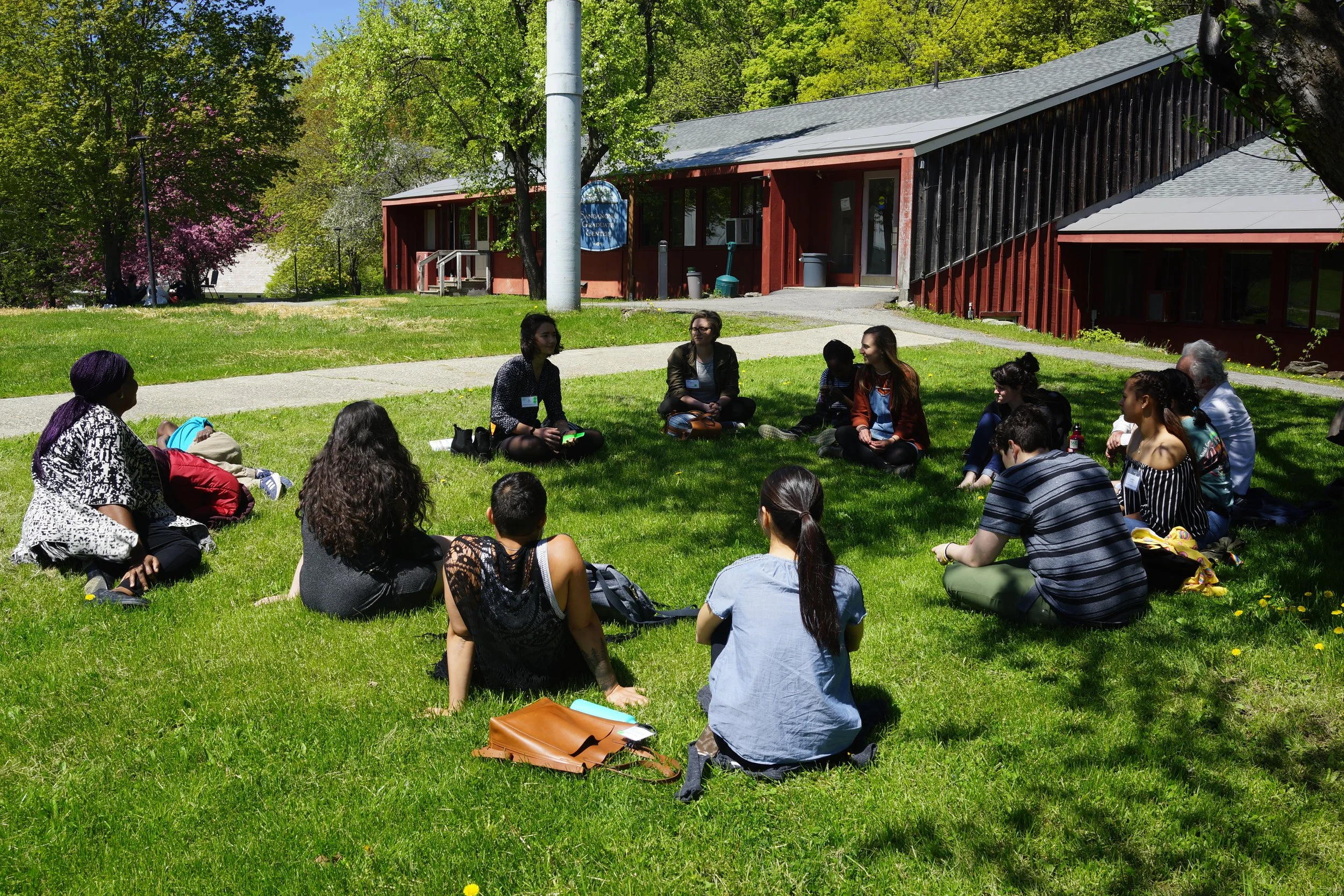 People sitting in a circle on green grass outside, engaged in a discussion or group activity, with trees and a building in the background.