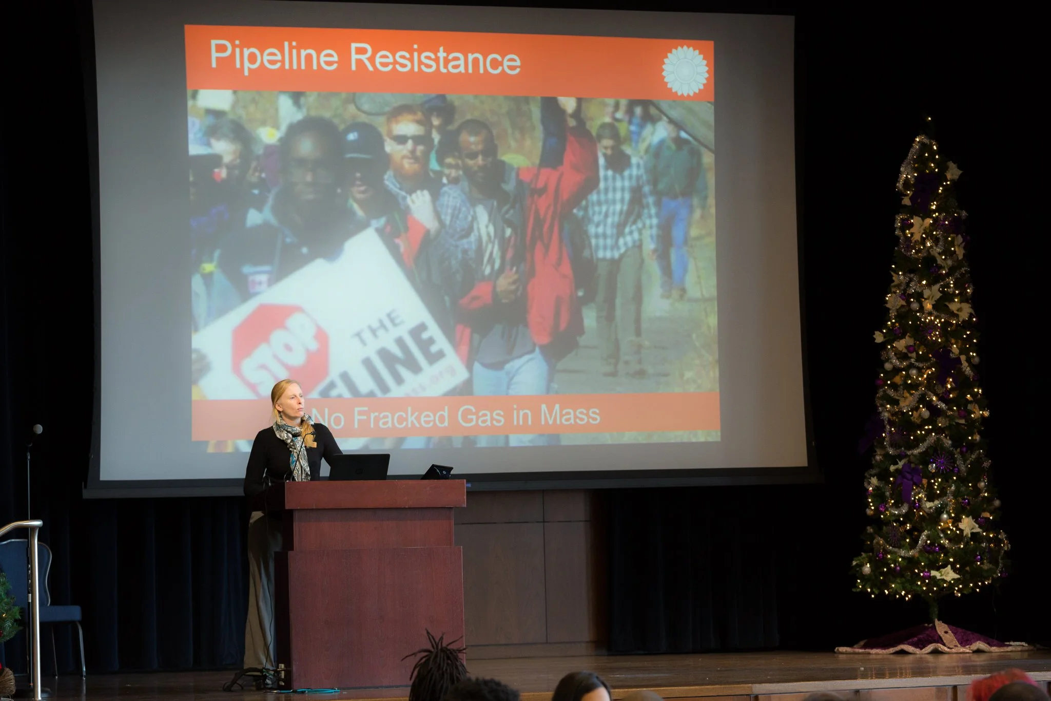A woman giving a presentation on a stage with a large screen behind her. The slide on the screen shows an image of people rallying, holding signs that say "Stop" and "No Fracked Gas in Mass," with the title "Pipeline Resistance" at the top. There is 