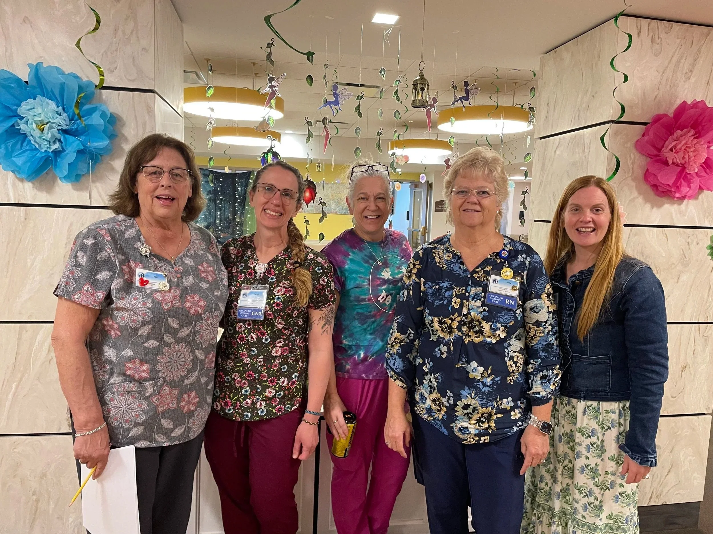 Five women standing in a hallway decorated with hanging fairy figures, large colorful paper flowers, and ribbon streamers, smiling at the camera.