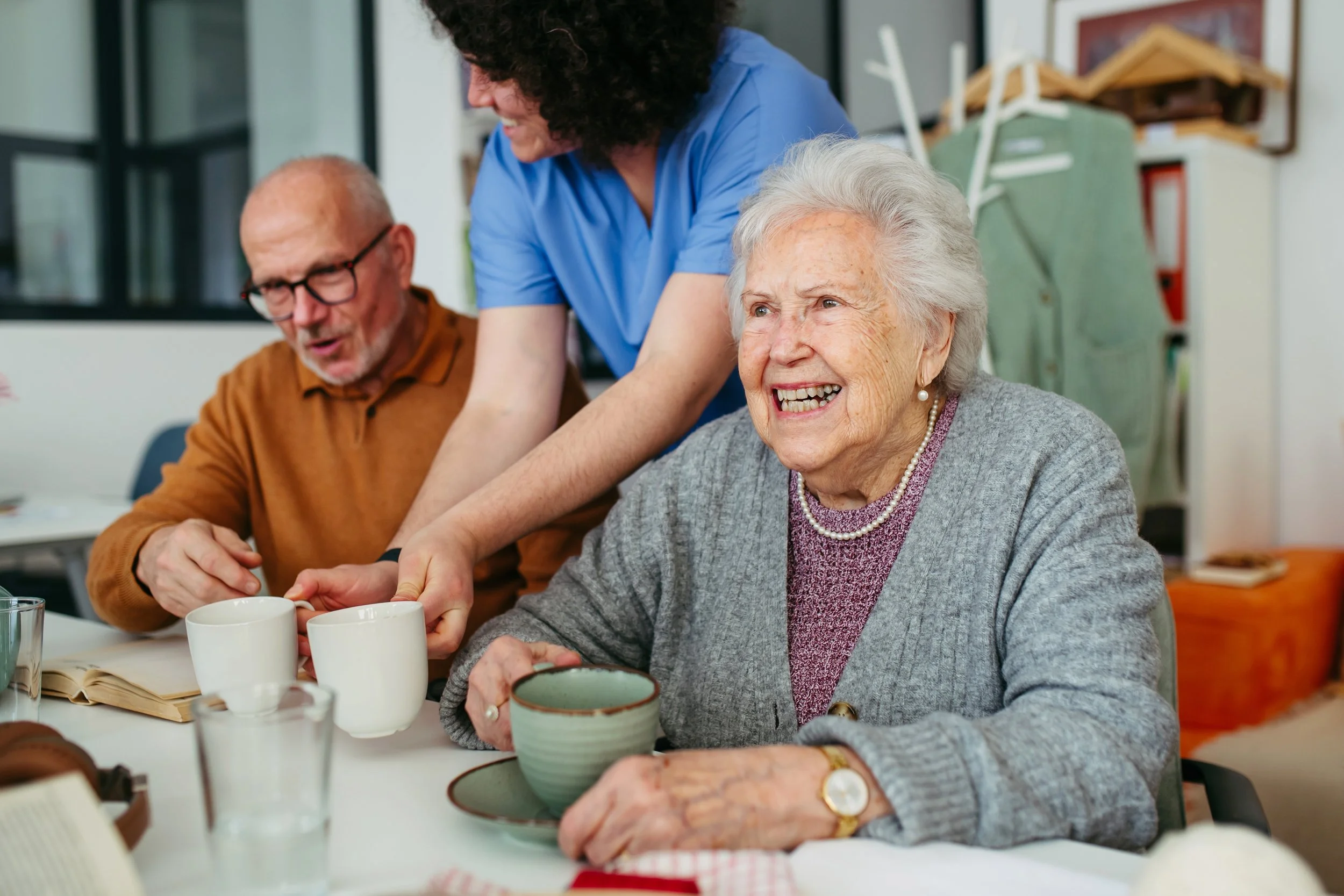 An elderly woman with gray hair and a gray cardigan smiling as she receives a cup of coffee from a caregiver. The caregiver, wearing blue scrubs, is leaning over and smiling at the elderly woman. There are cups, bowls, and glasses on the table in front of them, and a background of shelves and furniture.