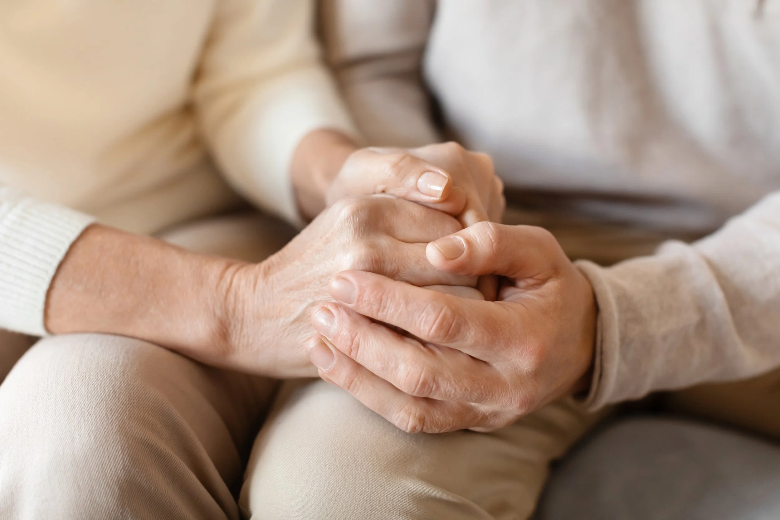 Close-up of two people holding hands, sitting closely together with beige clothing.