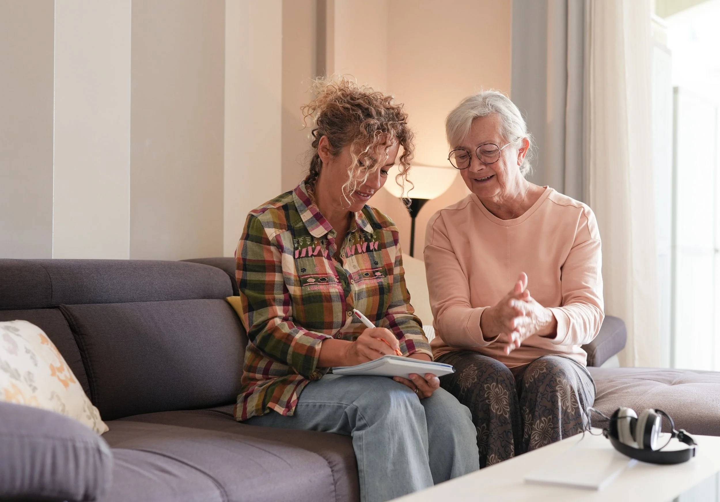 Two women sit on a sofa, one older and one younger, smiling and talking to each other. The older woman has white hair, glasses, and wears a pale pink top. The younger woman has curly hair, a colorful plaid shirt, and is holding a pen and notepad. They are in a well-lit living room with a lamp, curtains, and a window in the background.