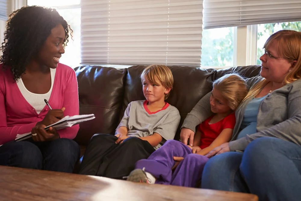 Therapist sitting on a couch with two children, a boy and a girl, and an adult woman, during a therapy session in a living room.