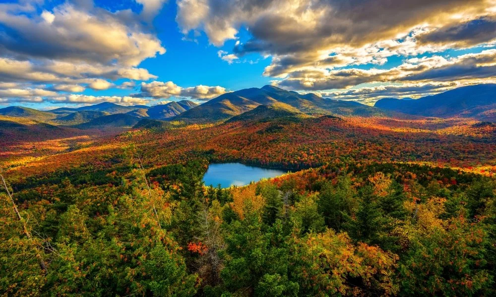 A vibrant autumn landscape of a mountain range with a small lake in the foreground, surrounded by colorful fall foliage and a partly cloudy sky.