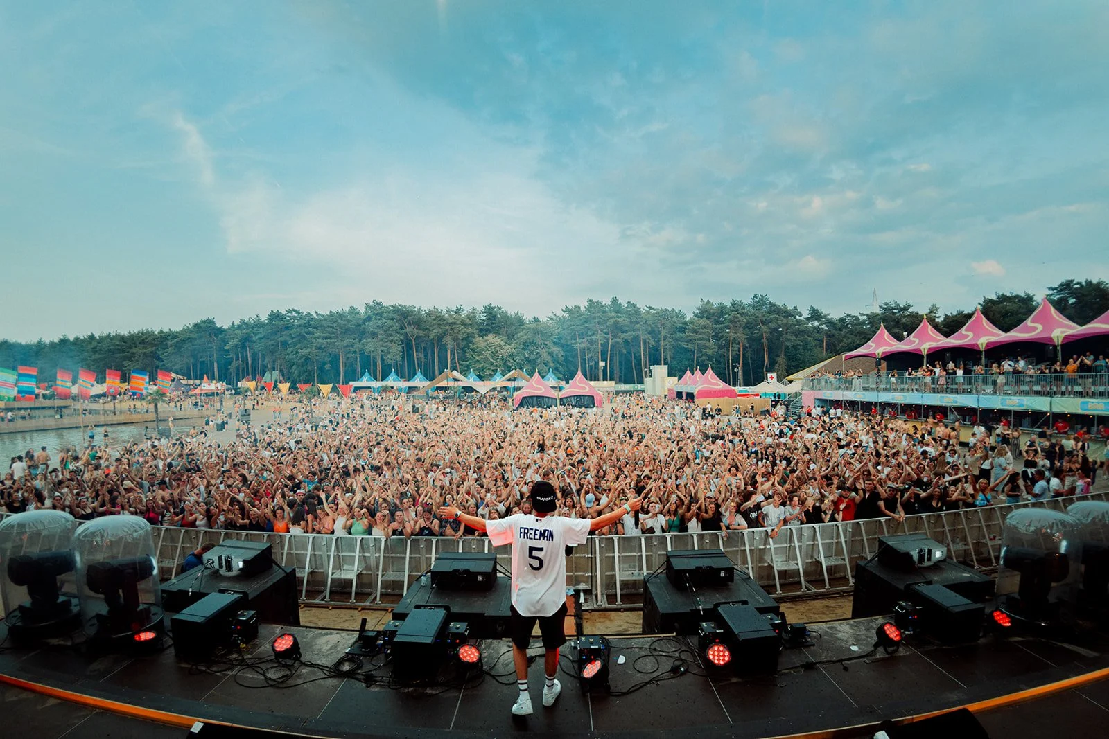 B2 Photography Jakob Hallaert  - Een DJ op een podium met uitgestrekte armen voor een groot publiek bij een openluchtfestival in de zomer, omringd door kleurrijke tenten en vlaggen, met een bos op de achtergrond.