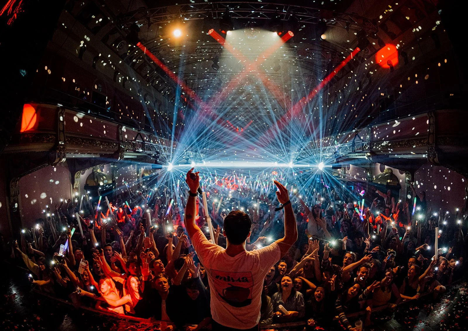 B2 Photography Jakob Hallaert  - Een groot dansfeest in een zaal met laserlichten en een enthousiaste menigte, de persoon op de voorgrond zwaait met beide handen omhoog.