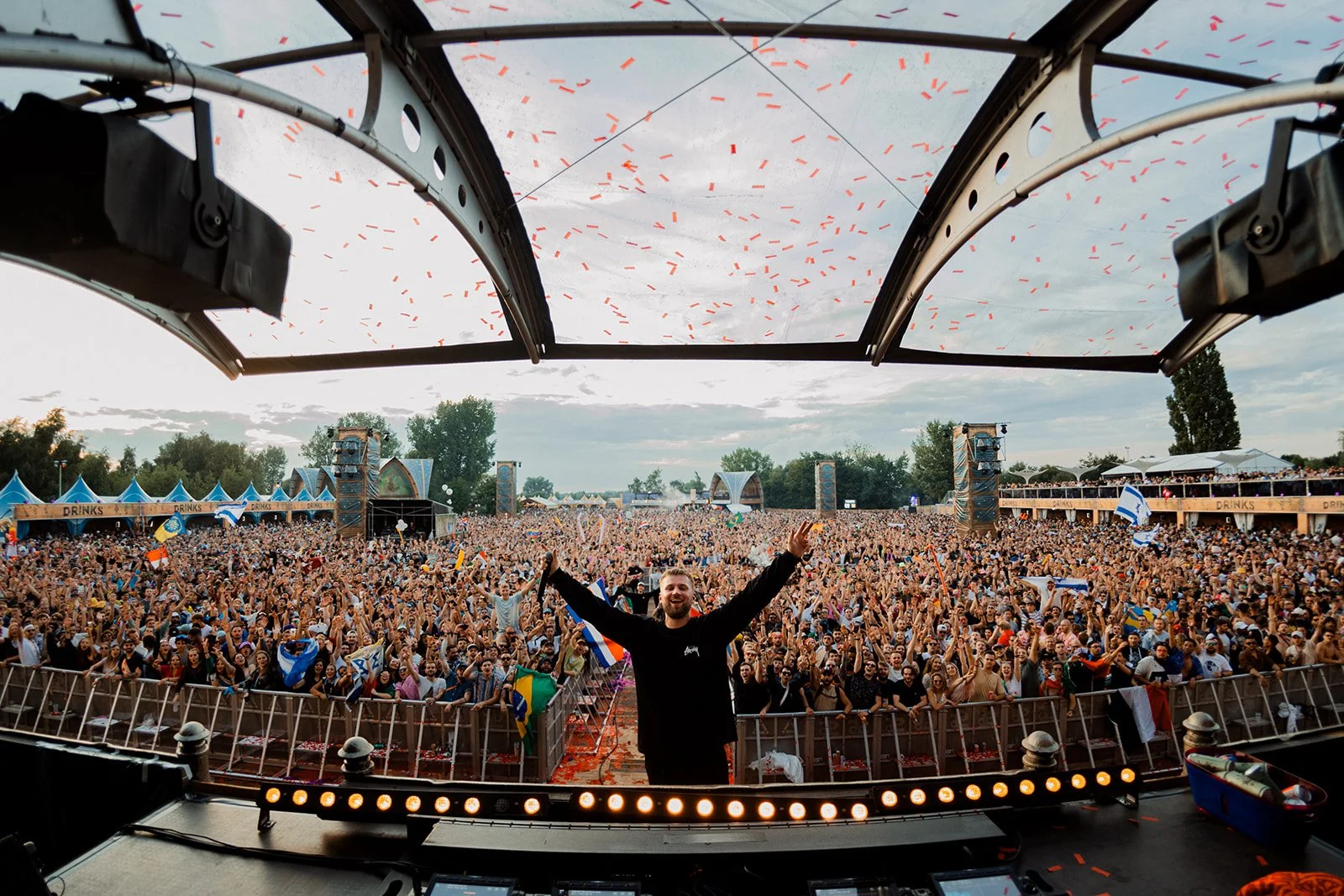 B2 Photography Jakob Hallaert  - Een DJ op een podium met een grote menigte tijdens een buitenfestival, feesthekken en vlaggen zichtbaar, confetti in de lucht, overdag met bewolkte hemel.