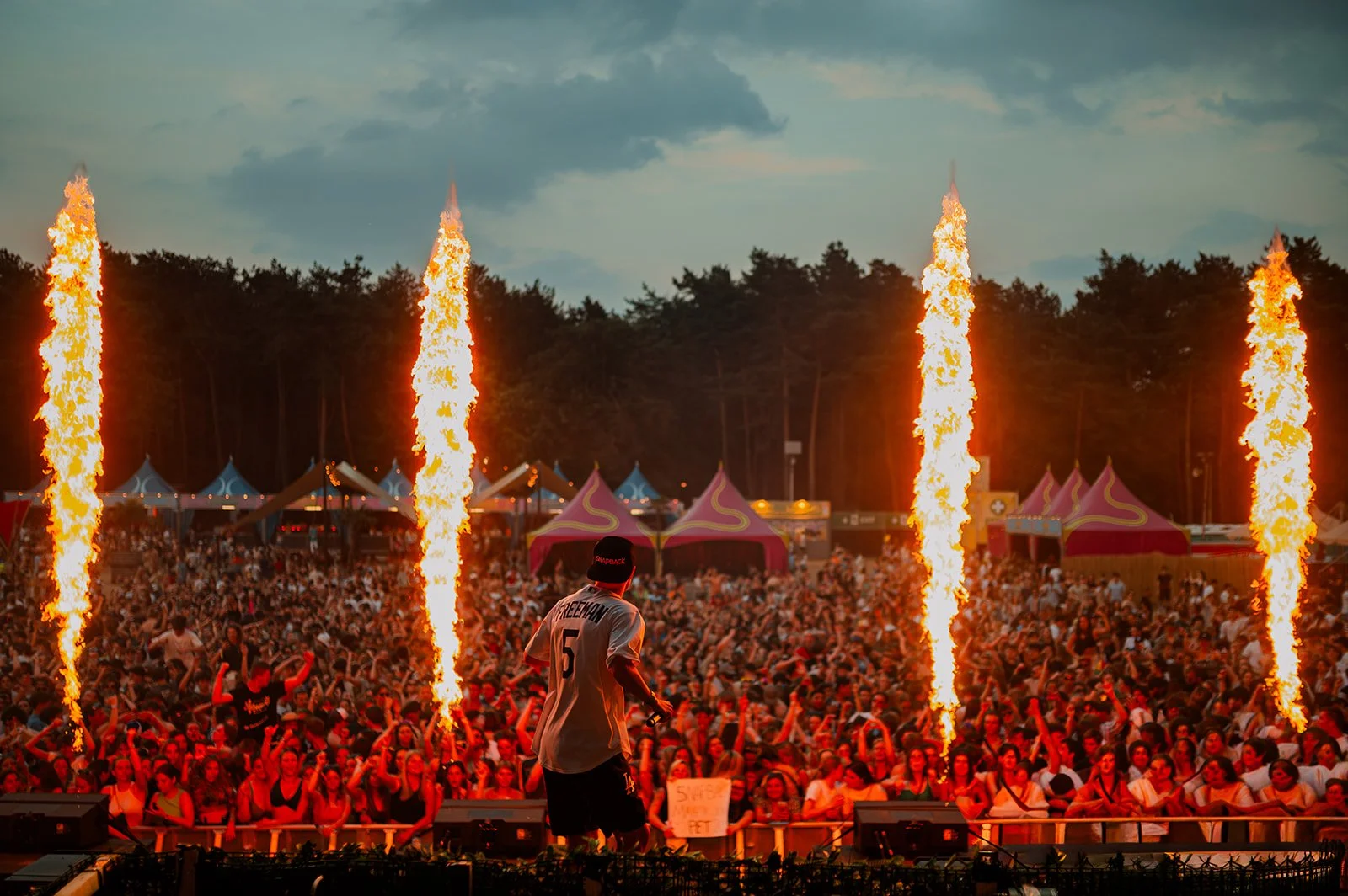 B2 Photography Jakob Hallaert  - Een druk muziekfestival met een groot publiek. Vlammen omhoog op het podium, een artiest met een pet en een sportshirt staat op het podium met zijn rug naar de camera, terwijl mensen in het publiek dansen en genieten.