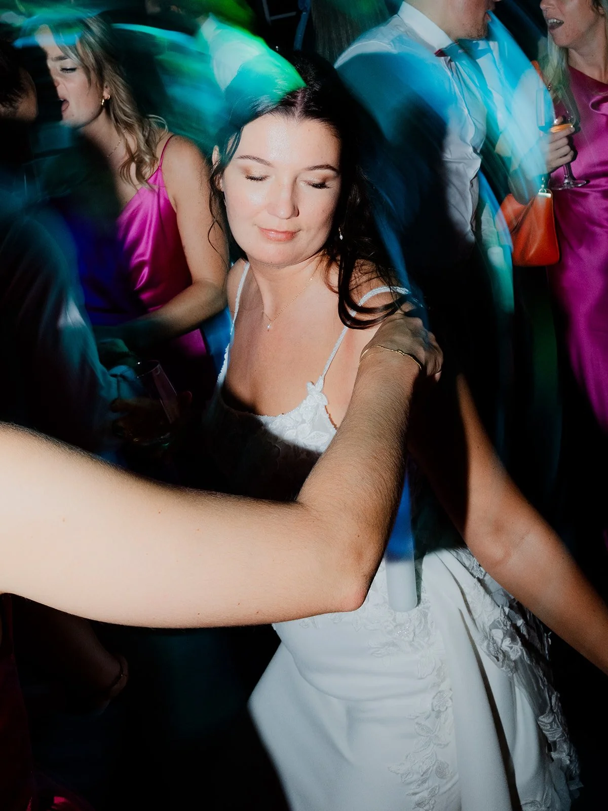 B2 Photography Jakob Hallaert  - Vrouw in witte jurk danst met haar ogen dicht op een feestje, omringd door mensen die plezier maken, met kleurrijke lichteffecten.