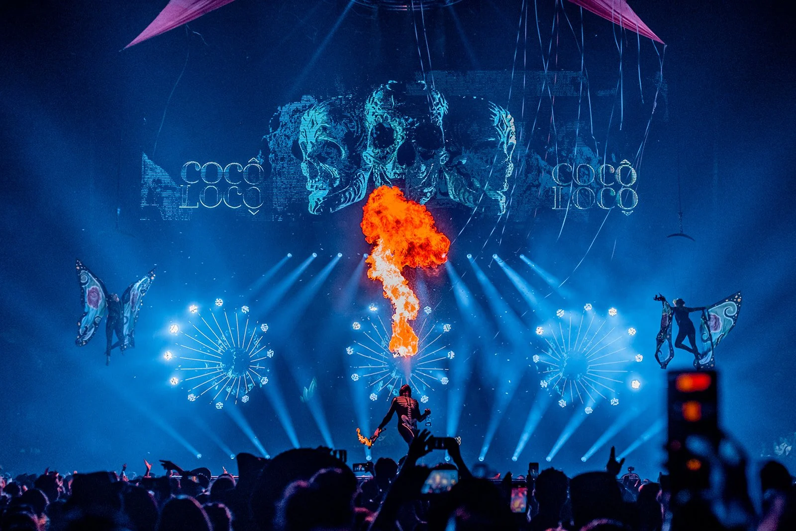 B2 Photography Jakob Hallaert  - Een nachtconcert met lichtshow, vuur, en dansers met vleugels op het podium, met een grote afbeelding van schedels op de achtergrond en het woord 'Cocoon' herhaald.