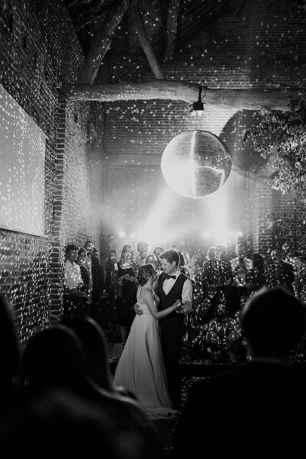 B2 Photography Jakob Hallaert  - Een zwart-wit foto van een bruiloftsdans, met een getrouwde paar dat in het midden danst, omringd door gasten in een binnenruimte met stenen muren en een disco-bal die het licht verspreidt.