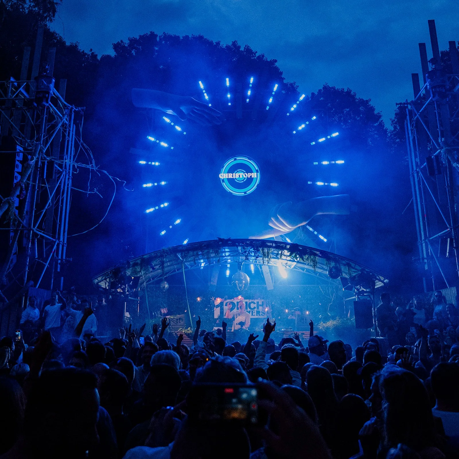 B2 Photography Jakob Hallaert  - Een menigte mensen bij een openlucht muziekfestival met een verlichte podium en een DJ, onder een nachtelijke hemel met blauwe lichten.