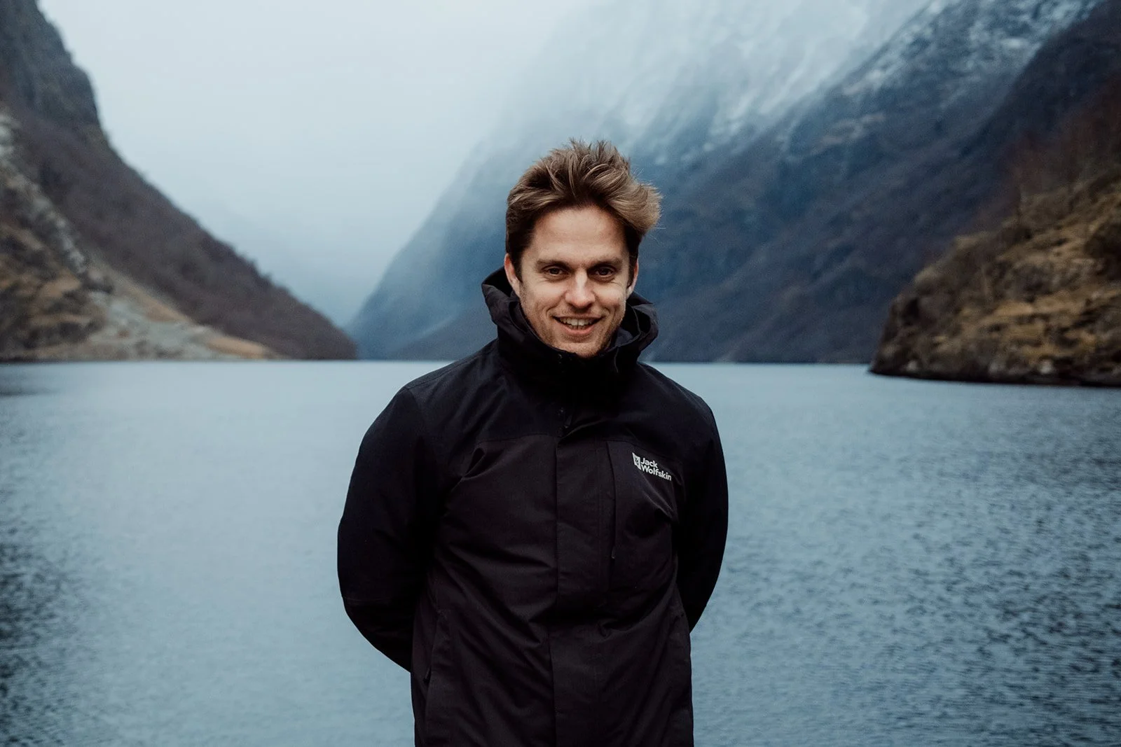 B2 Photography Jakob Hallaert  - Man met zwart Jack Wolfskin jas, lachend, voor een fjord met bergen, water en mist op de achtergrond