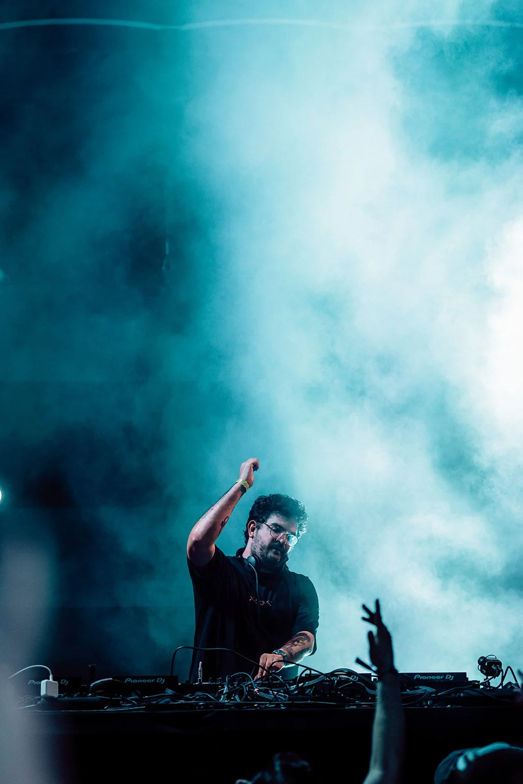 B2 Photography Jakob Hallaert  - Een man achter een dj-set tijdens een live muziekoptreden, omringd door blauwe en groene rook, met mensen in het publiek die naar hem kijken en hun handen omhoog stekken.