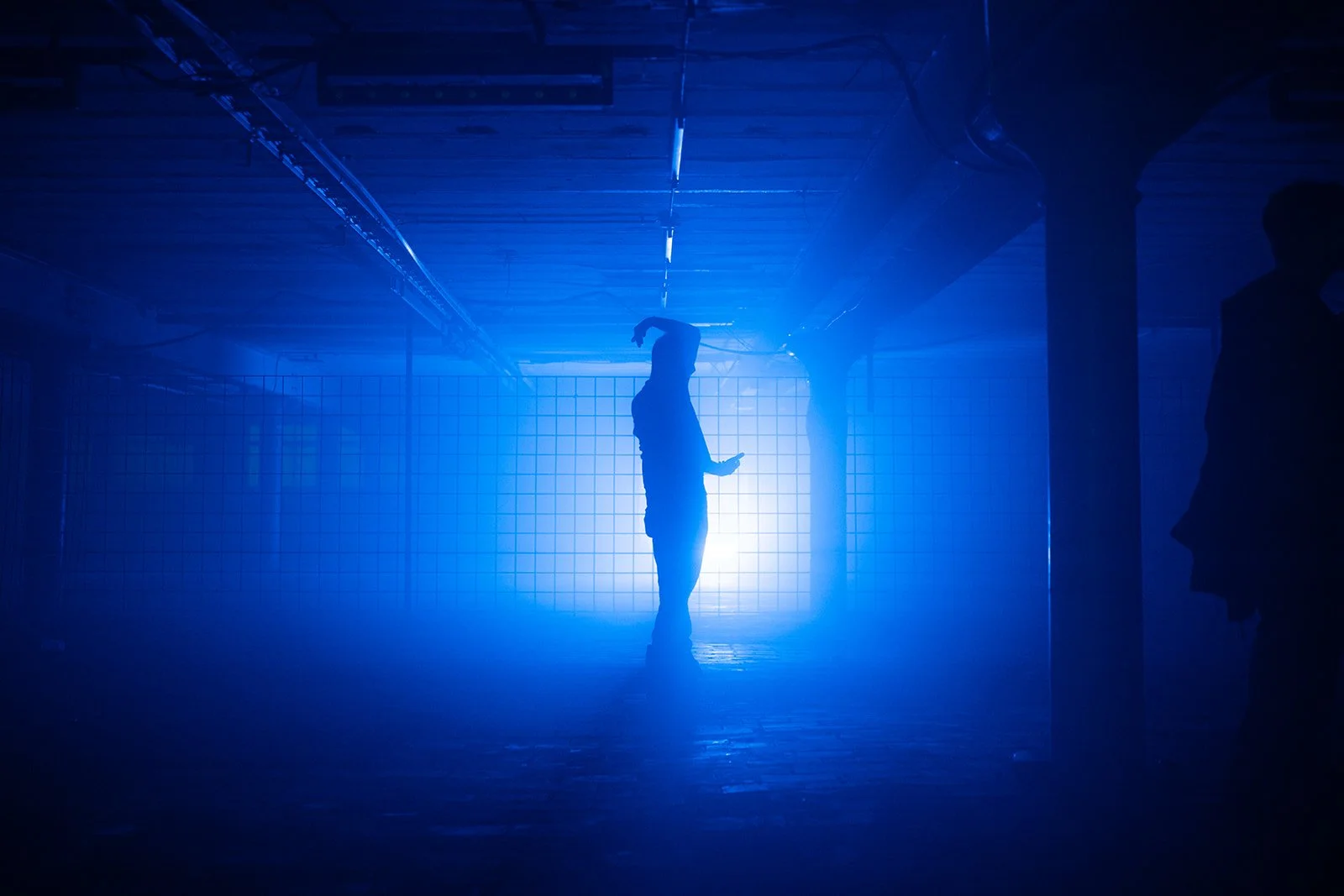B2 Photography Jakob Hallaert  -  silhouet van een persoon die dans of poses in een kamer met blauwe verlichting, omringd door een rooster muur en ondersteunende stutten.