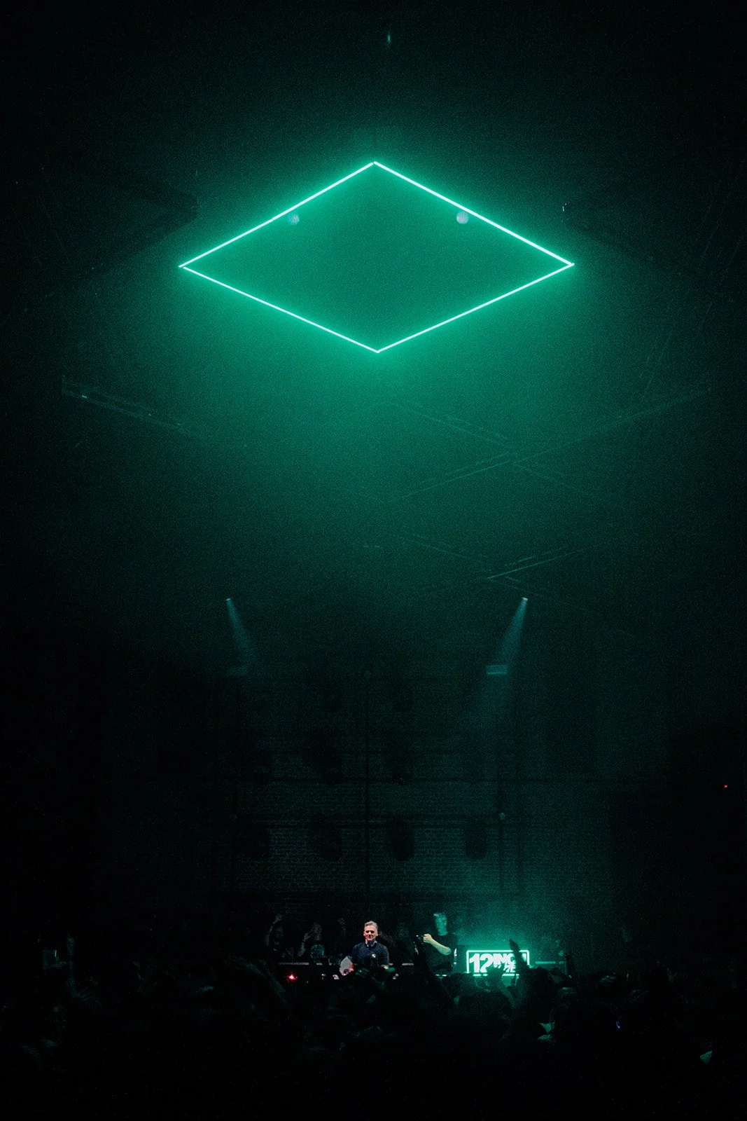 B2 Photography Jakob Hallaert  - Een donkere nachtclub met een DJ die achter de draaitafels staat, verlicht door een grote groene neonlicht in de vorm van een vierkant. Bezoekers dansen in de duisternis.