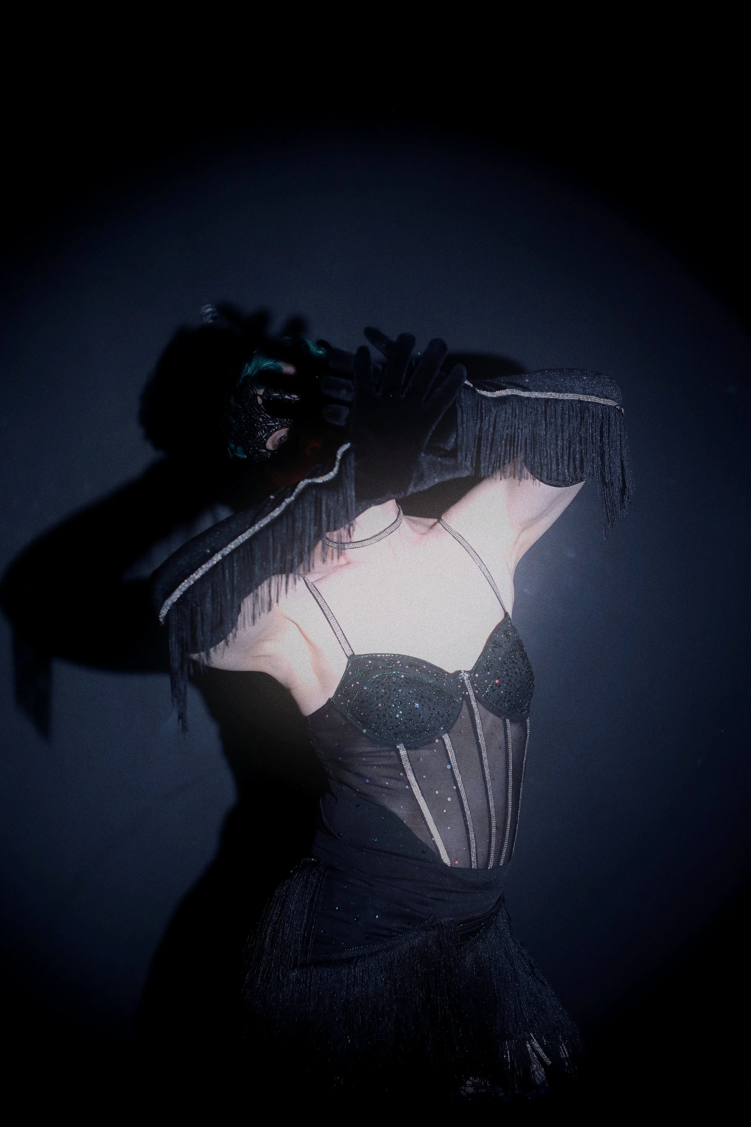 A person in a theatrical costume, dressed in shiny black fabric with fringe details, wearing a headdress and black gloves, on a dark stage.