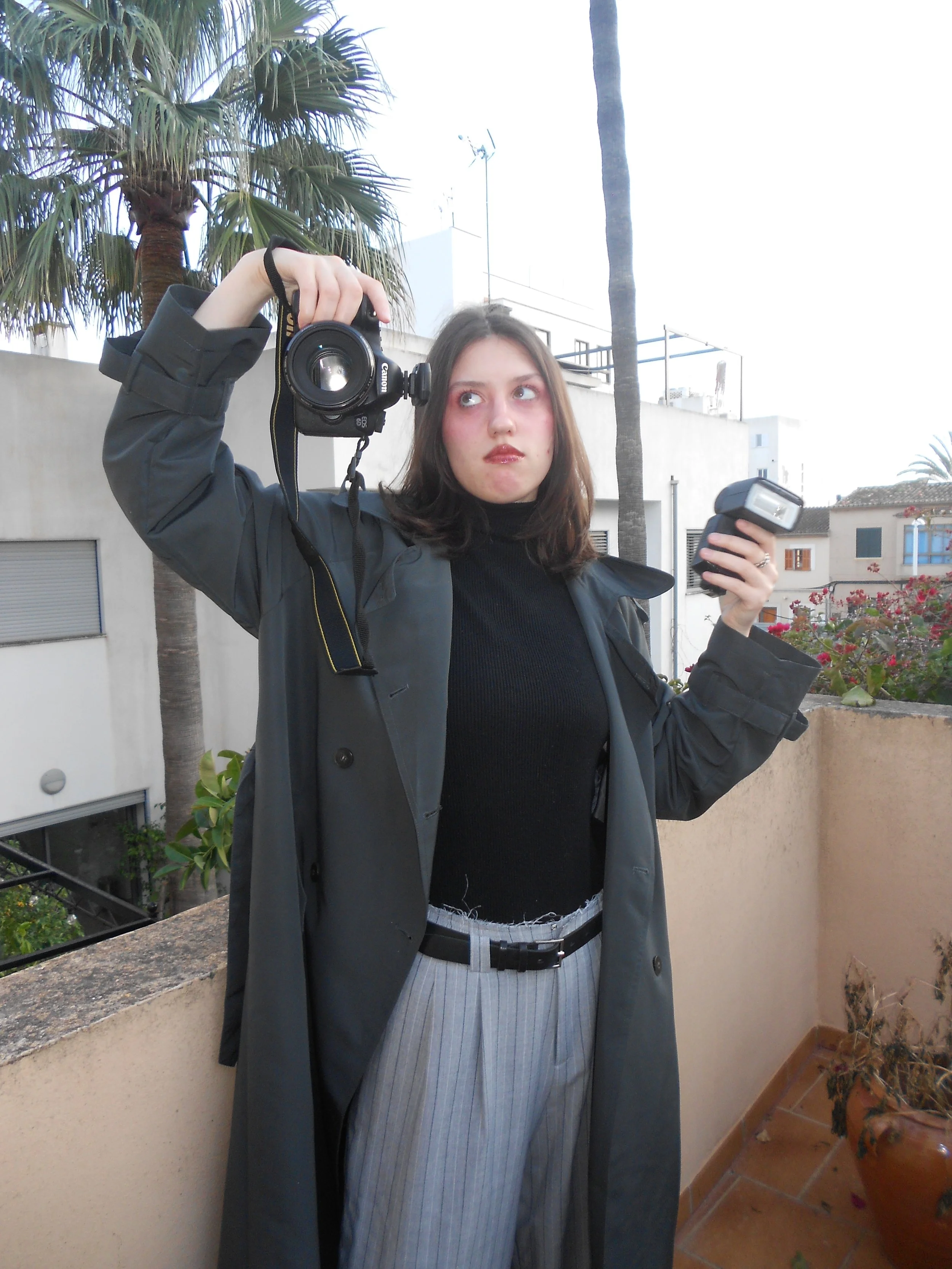 Young woman in a dark jacket, black shirt and striped pants, holding a DSLR camera and flash, on a balcony with plants and buildings in the background.