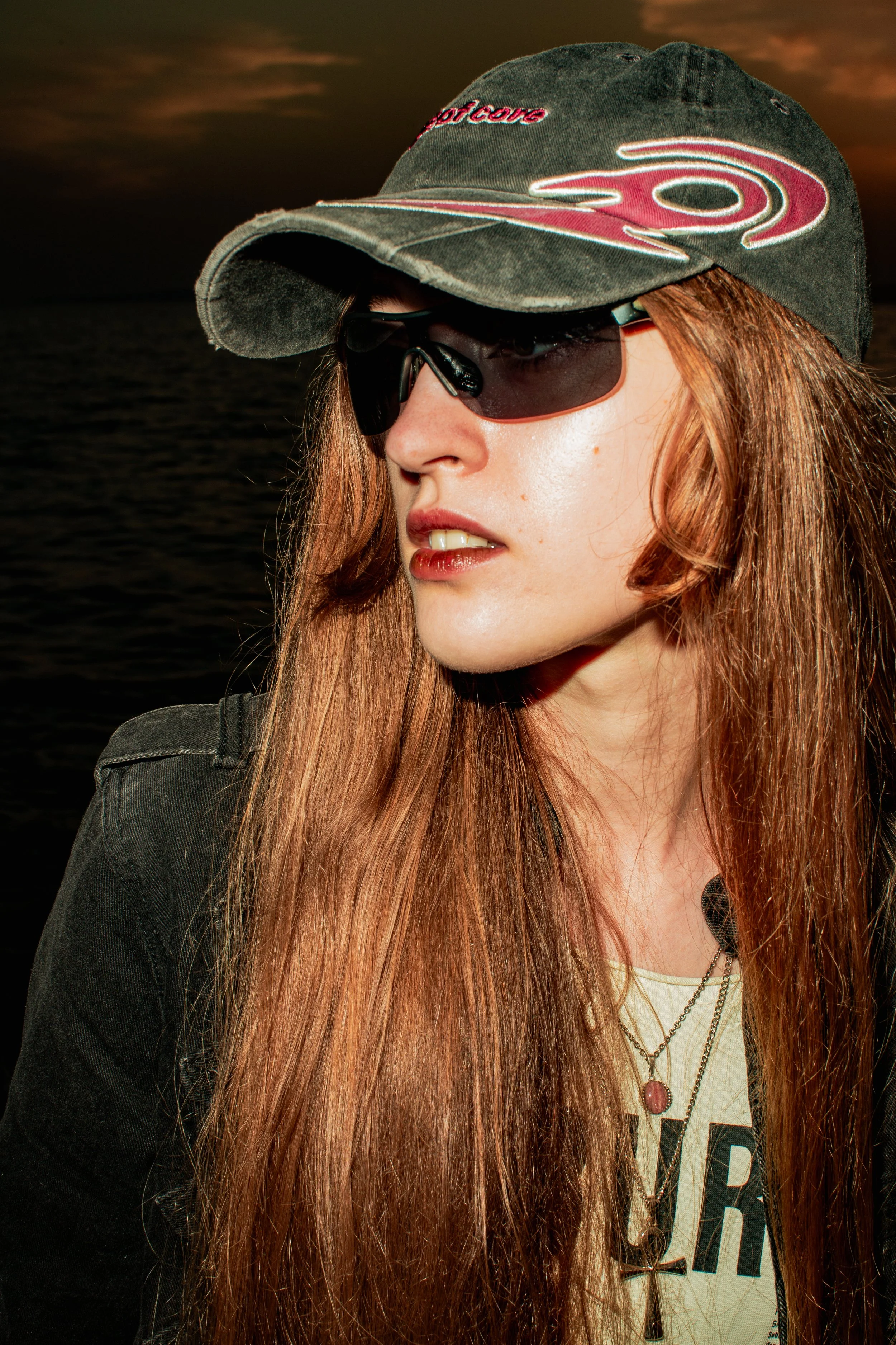 Portrait of a young woman with long, curly hair, wearing black sunglasses, a green cap with embroidery and a white shirt with a graphic and several chains, against a dark background with a cloudy sky.