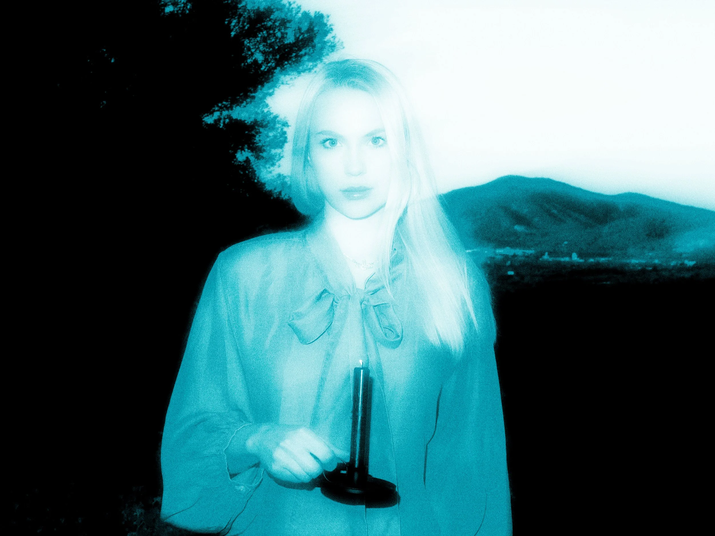 A woman with long, blonde hair, wearing a light-colored shirt and scarf, holds a microphone. In the background, hills and trees can be seen under a clear sky.